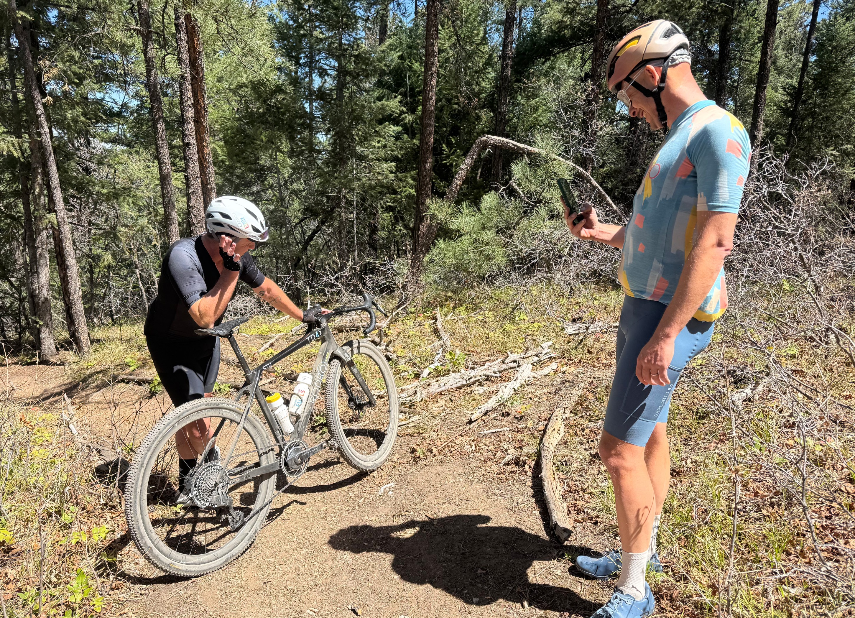 Two cyclists taking a break on a trail in a wooded area, with one checking their phone and the other leaning on a bike.