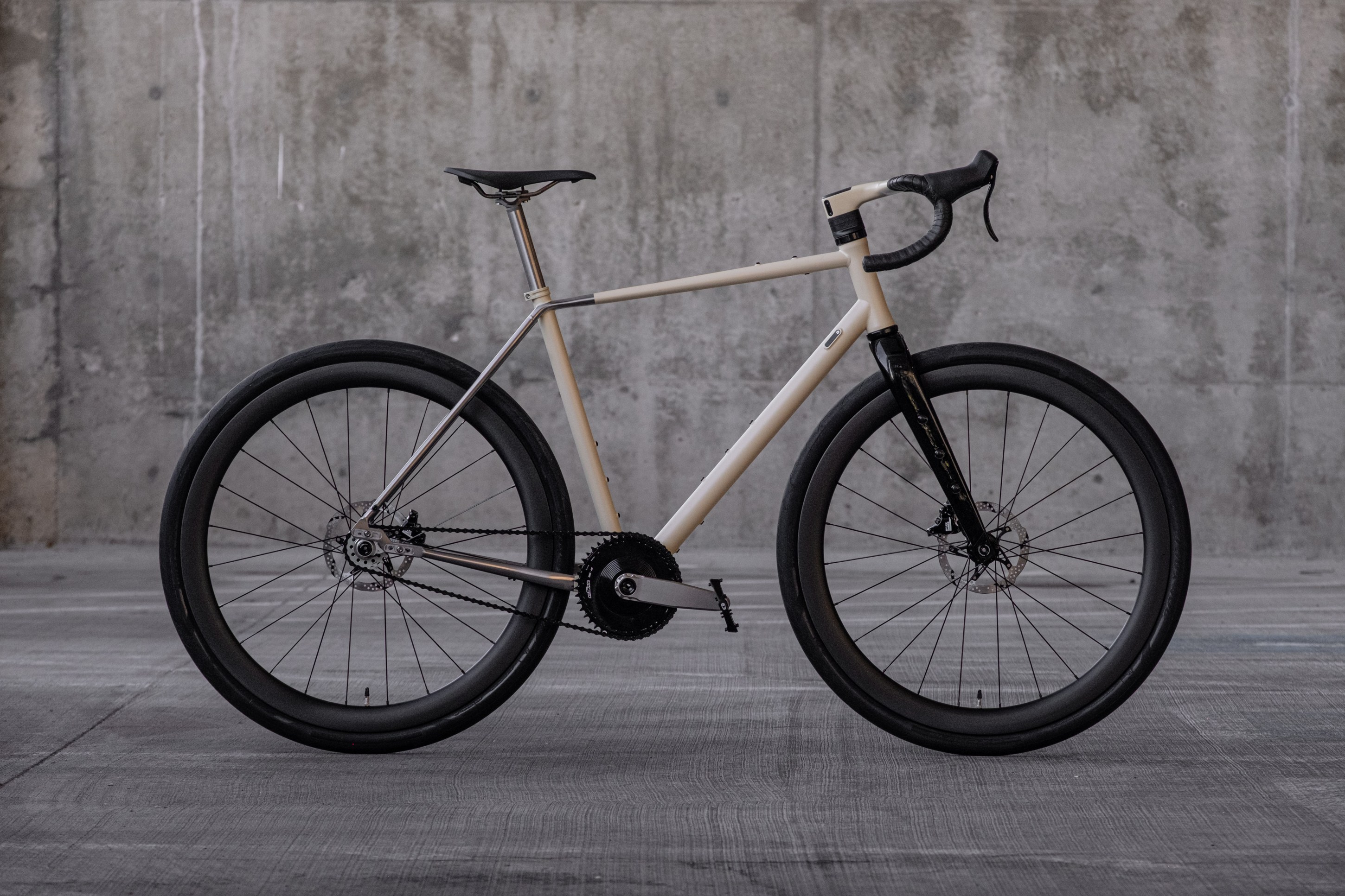A minimalist bicycle with a sleek design, featuring a beige frame and black wheels, positioned against a concrete background.