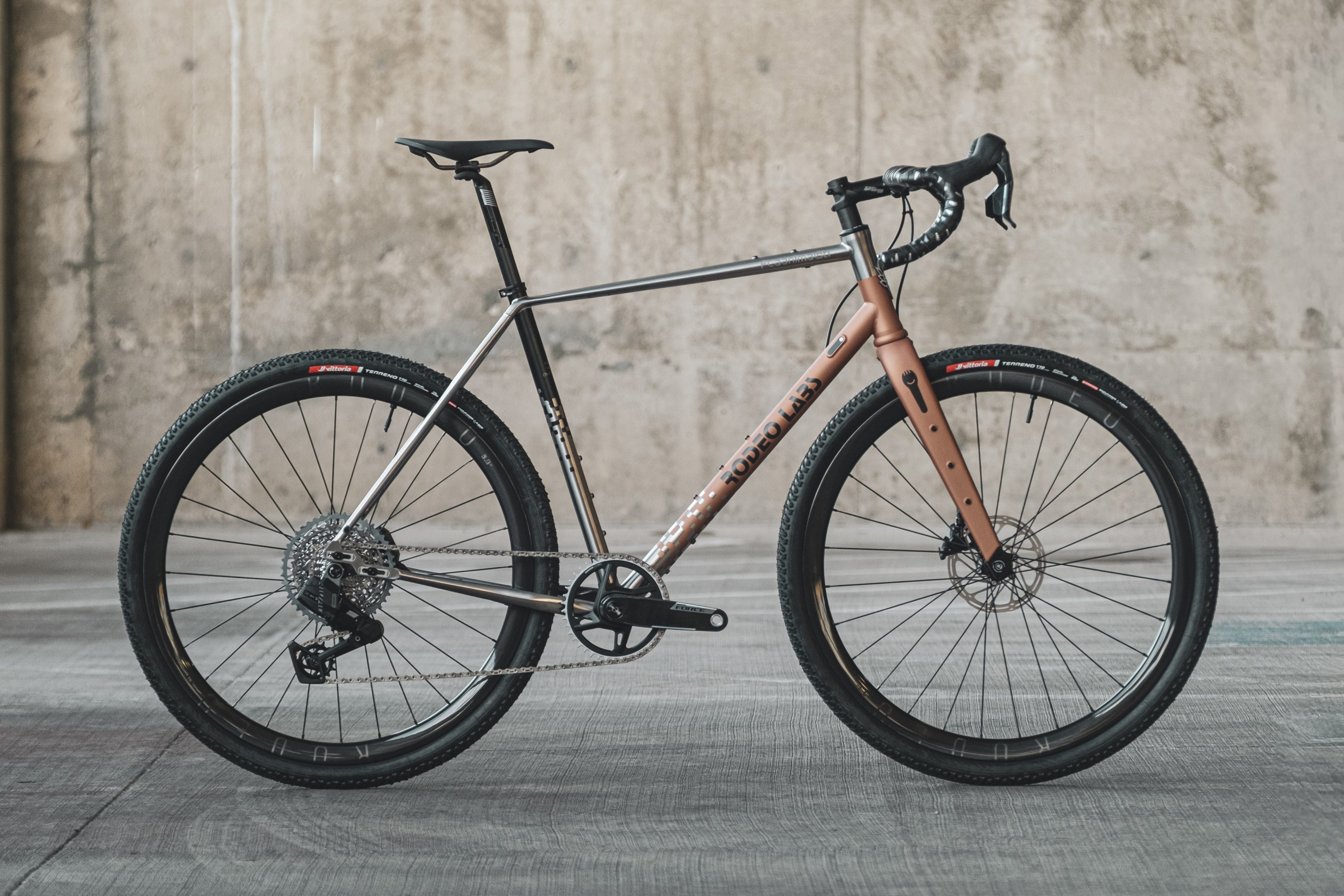 A modern gravel bike with a sleek design, featuring a titanium frame and copper accents, positioned against a concrete background.