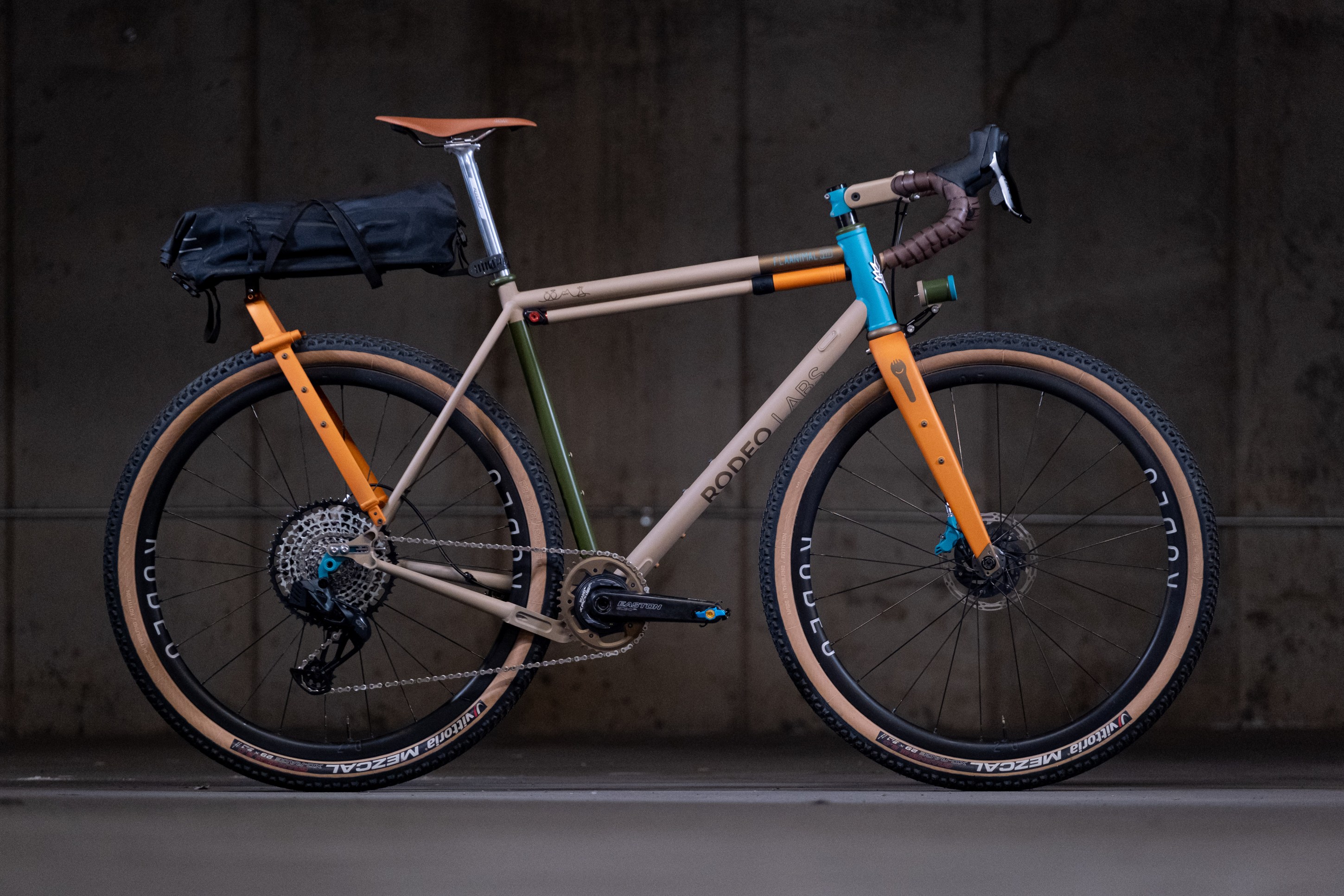 A stylish bicycle with a unique color scheme, featuring a beige frame with orange, green, and blue accents. It has a black bag mounted on the rear and wide tires suitable for gravel riding.