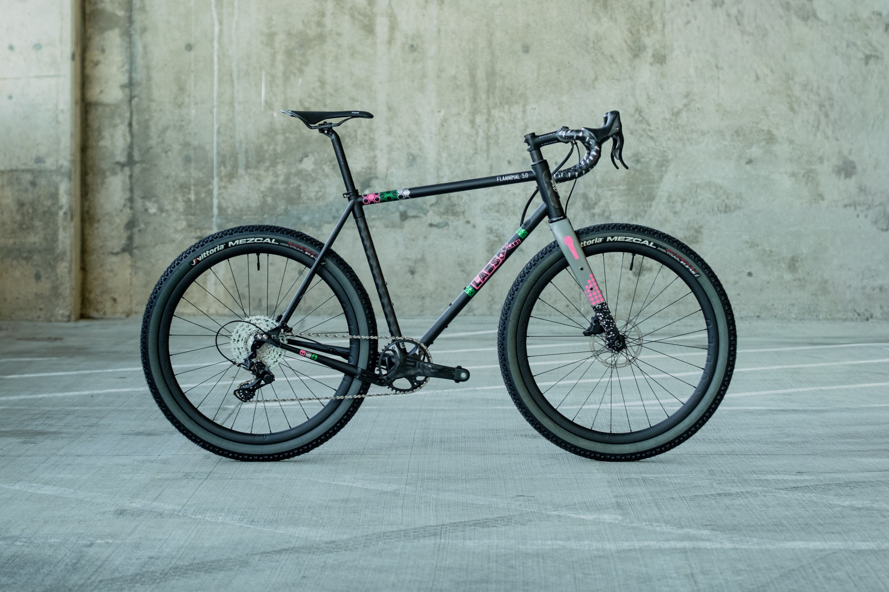 A black gravel bike with colorful accents, positioned against a gray concrete wall in a parking area.