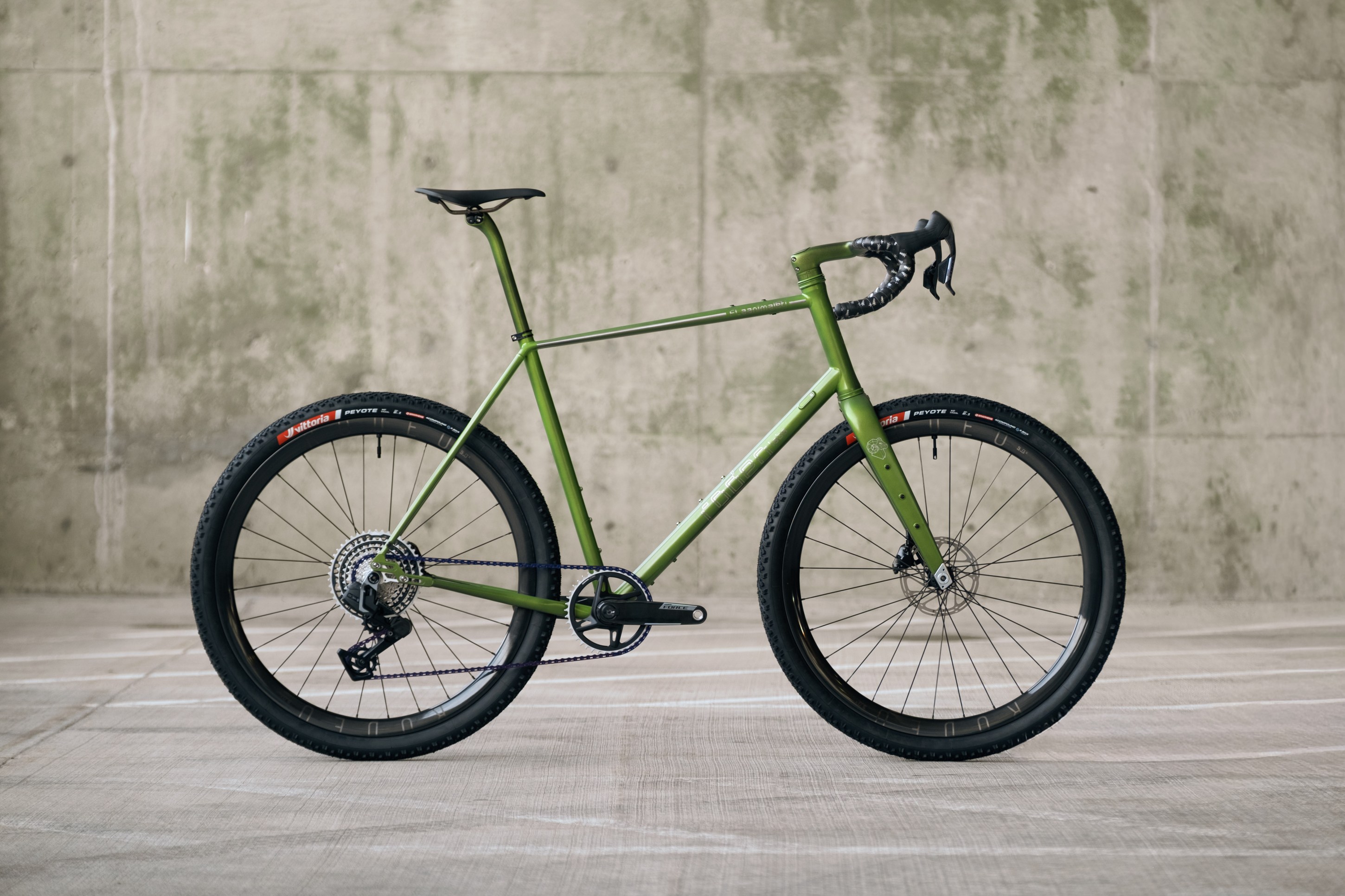A green gravel bike displayed against a textured concrete wall, showcasing its sleek frame, drop handlebars, and wide tires.