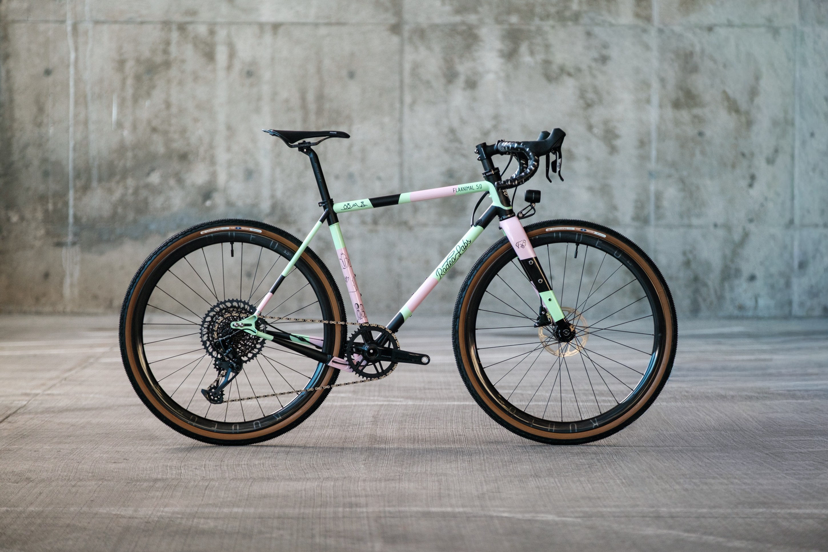 A colorful bicycle with a mint green and pink frame, featuring black handlebars and wheels, displayed against a gray concrete background.