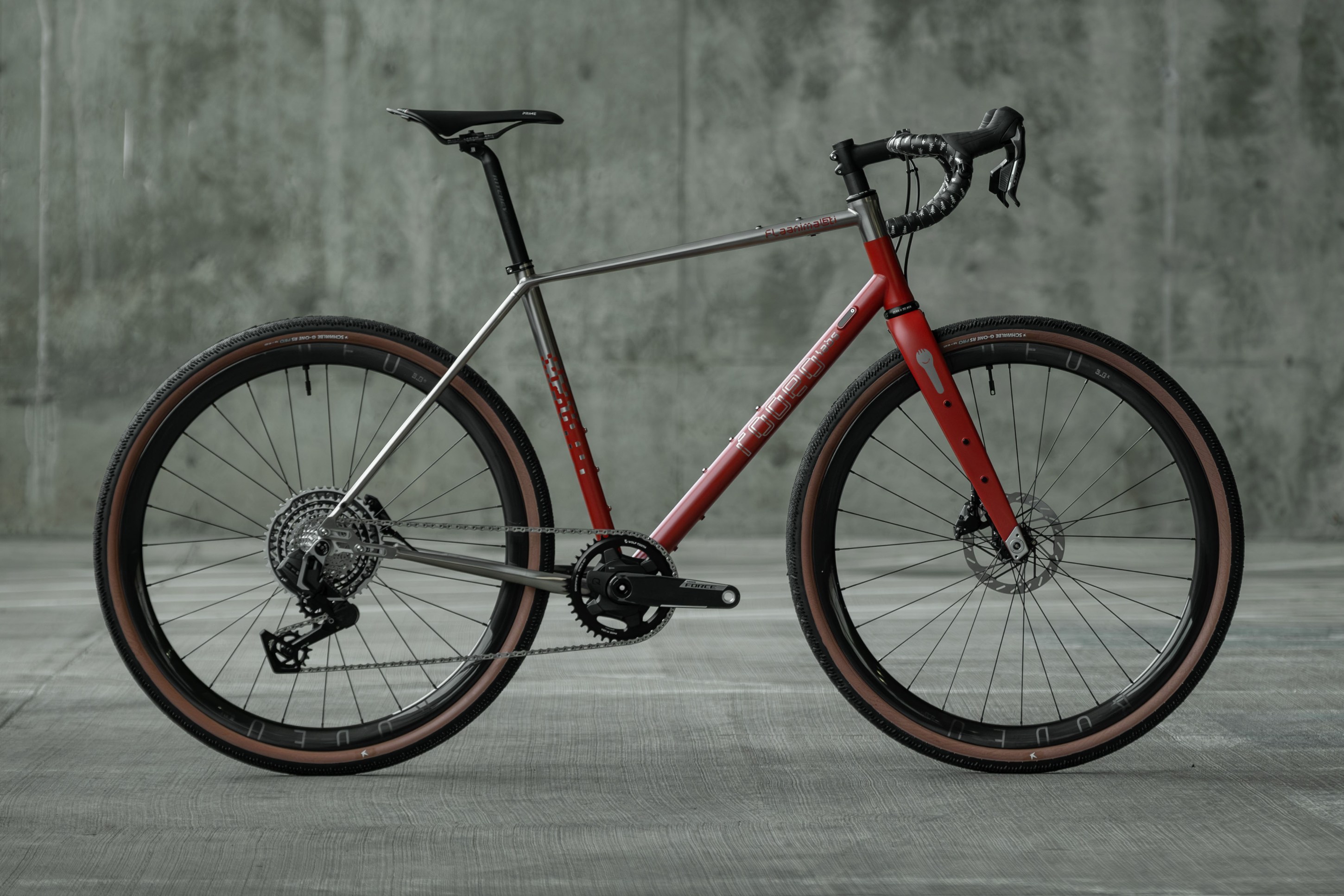 A sleek, modern bicycle with a titanium and red frame, featuring black handlebars, disc brakes, and brown tires, set against a concrete backdrop.