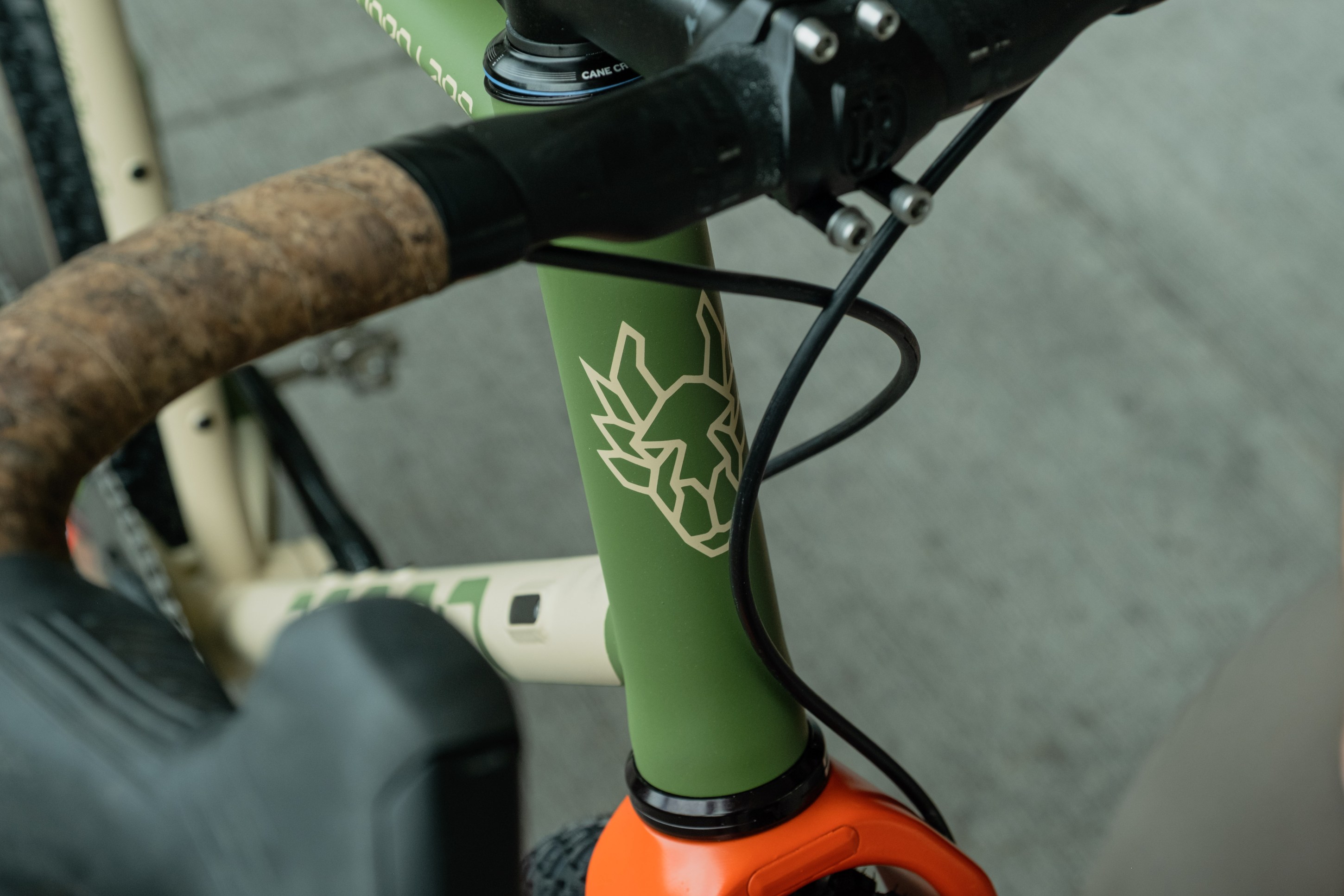 Close-up view of a bicycle frame featuring a green finish with a stylized animal logo, orange fork, and cork handlebar grip.