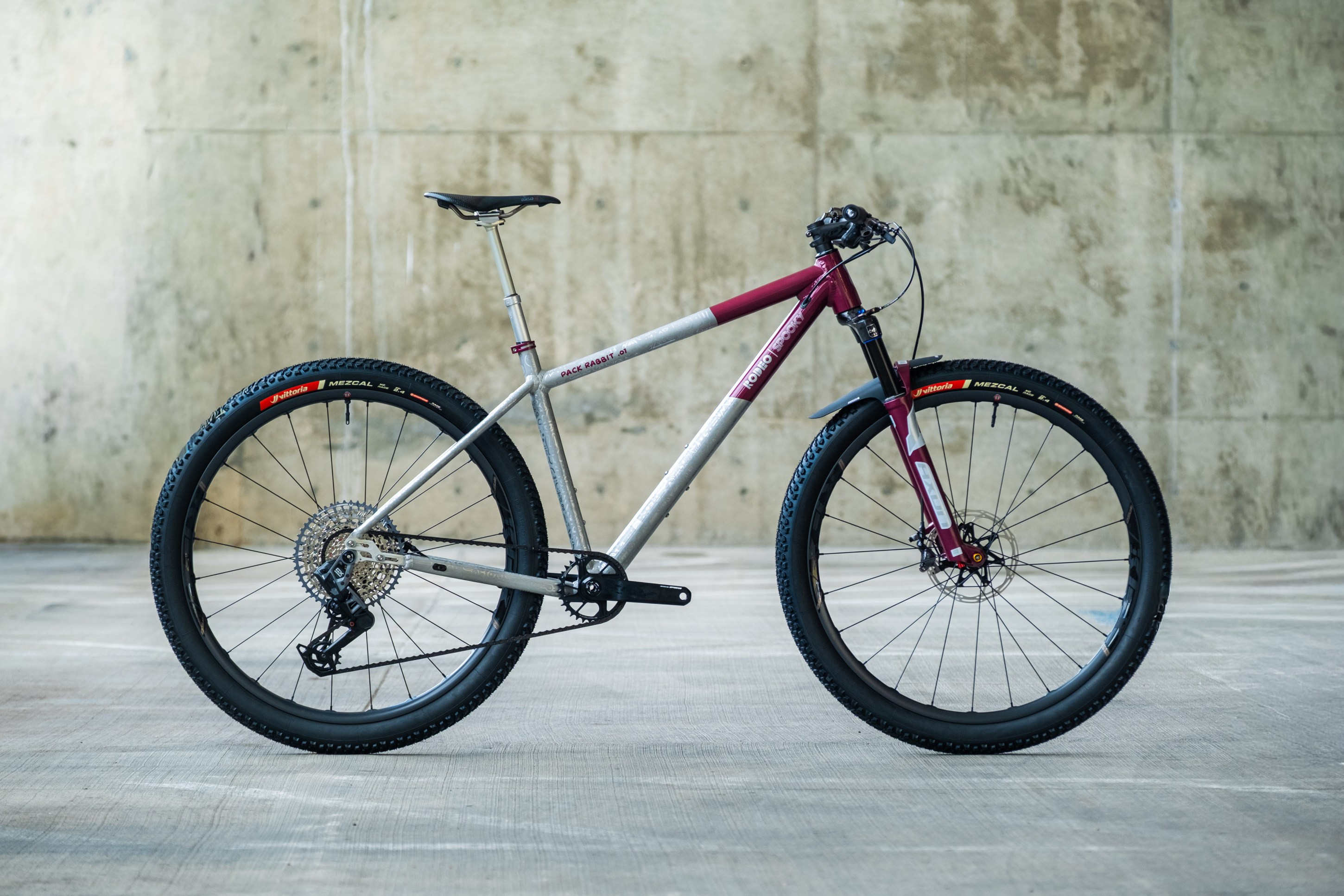 A high-quality mountain bike with a silver and maroon frame, featuring wide tires and a sleek design, positioned against a textured concrete background.