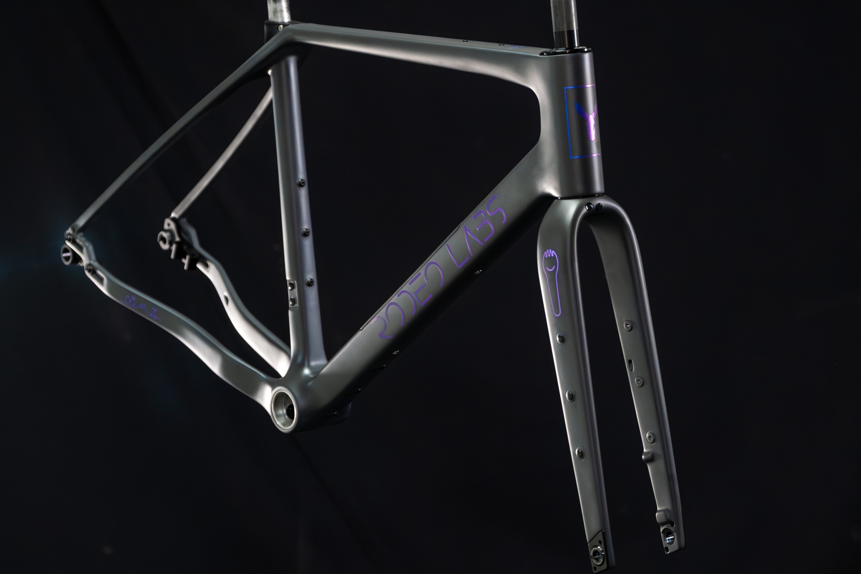 A close-up of a modern bicycle frame featuring a matte metallic finish and purple branding, suspended against a dark background.