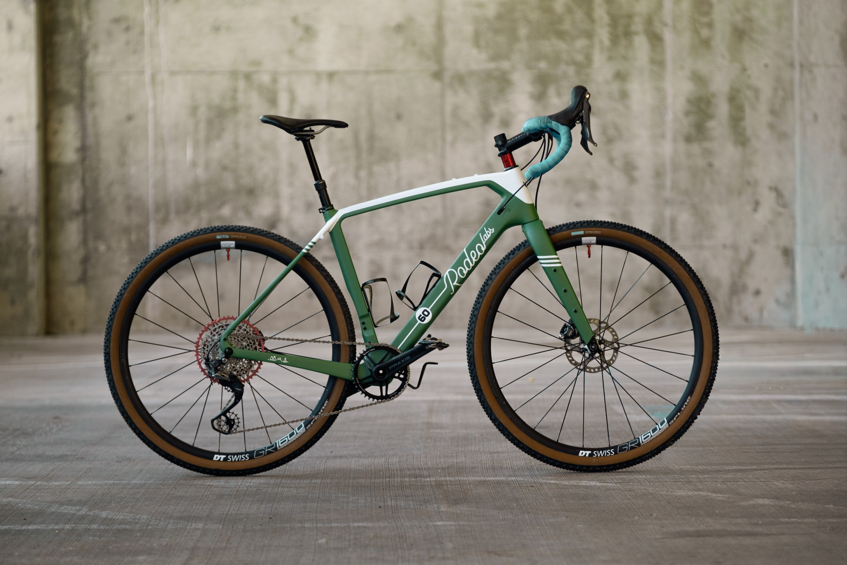 A green gravel bike with a sleek design, featuring thick tires and disc brakes, positioned against a textured concrete background.