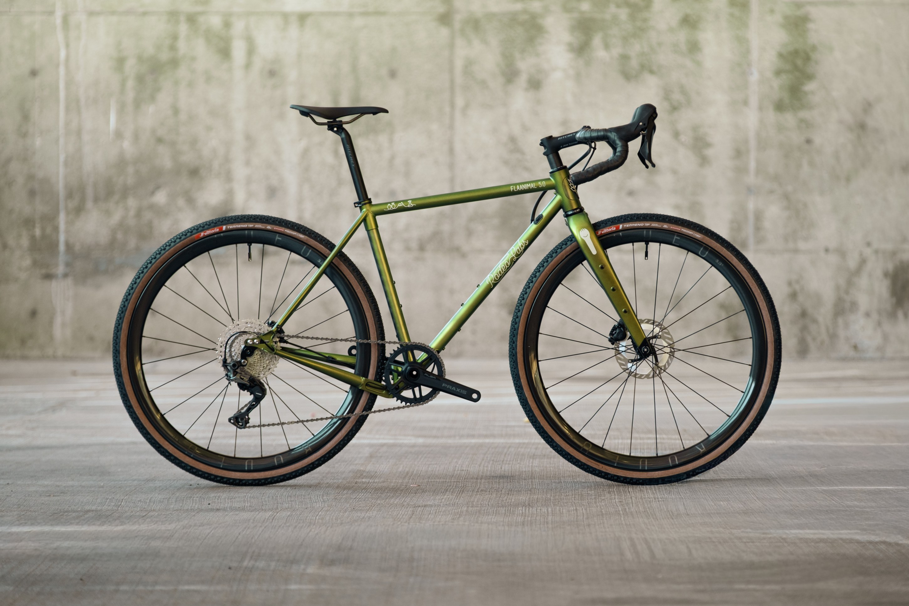 A green gravel bike with disc brakes and knobby tires, positioned sideways against a textured concrete backdrop.