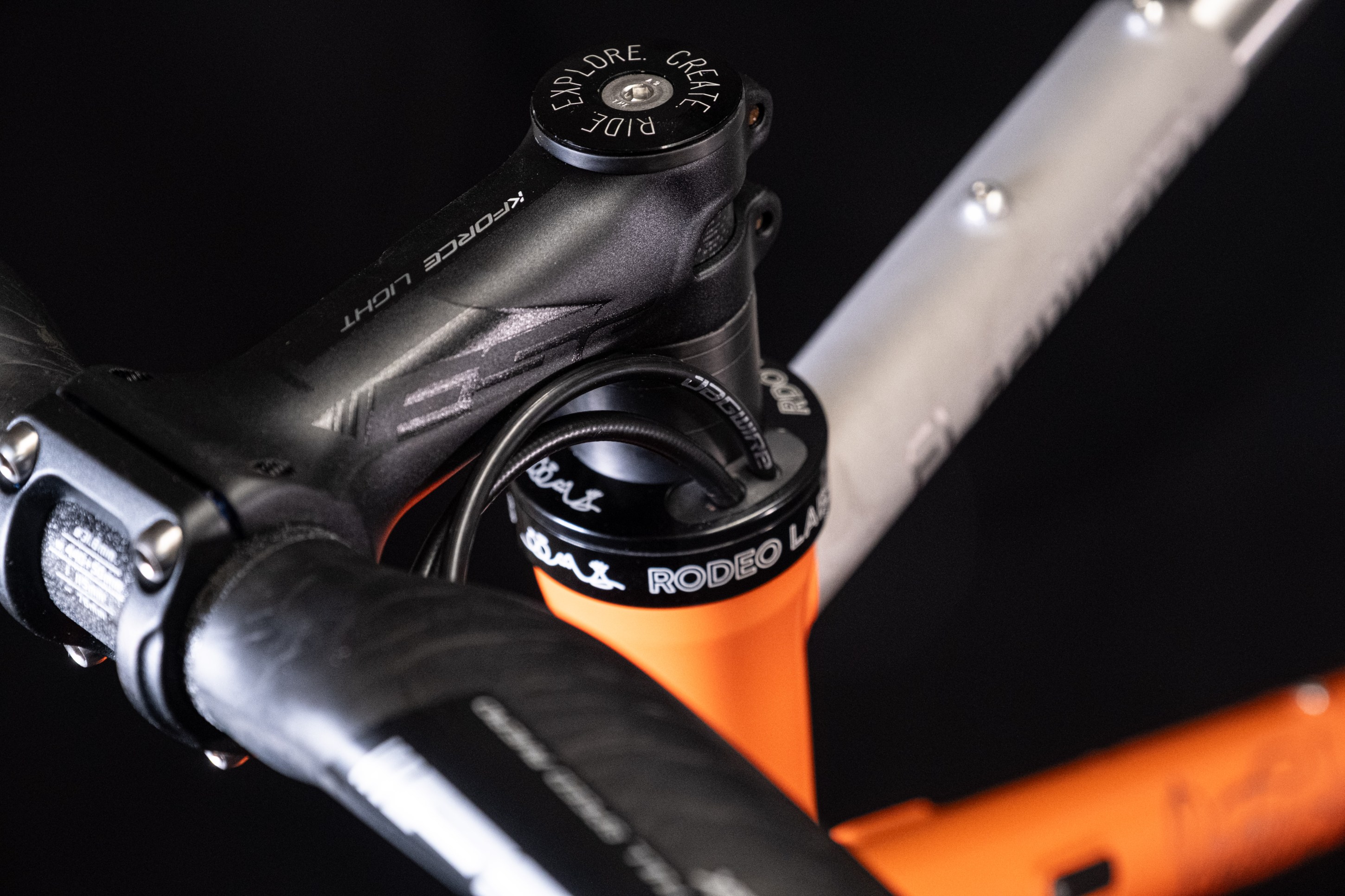 Close-up of a bicycle's handlebar stem and headset, showcasing black and orange components with the text 'Rodeo Labs' and 'Explore. Create. Ride.'