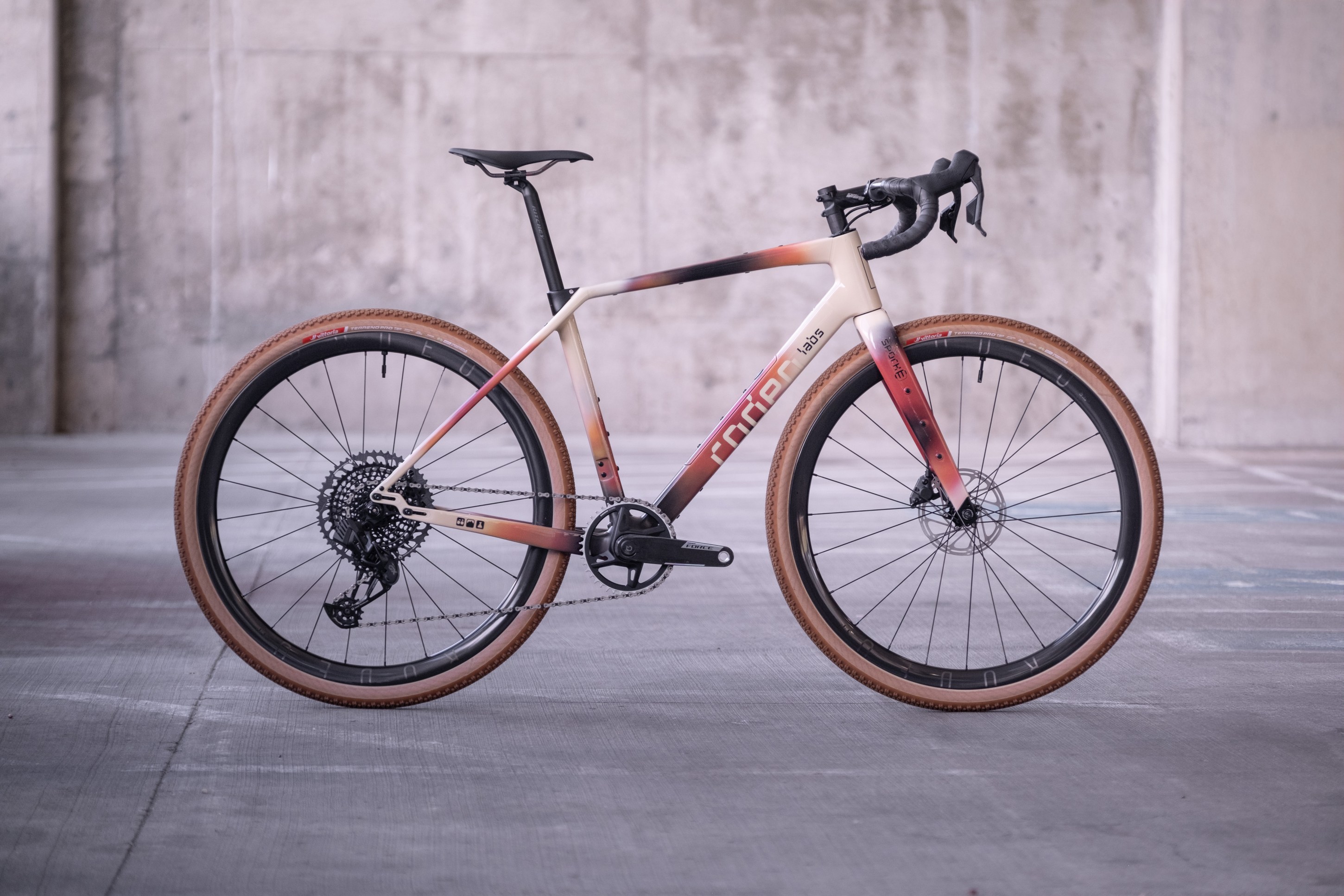 A modern gravel bike with a sleek, multicolored frame and knobby tires, parked in an industrial space with a concrete backdrop.