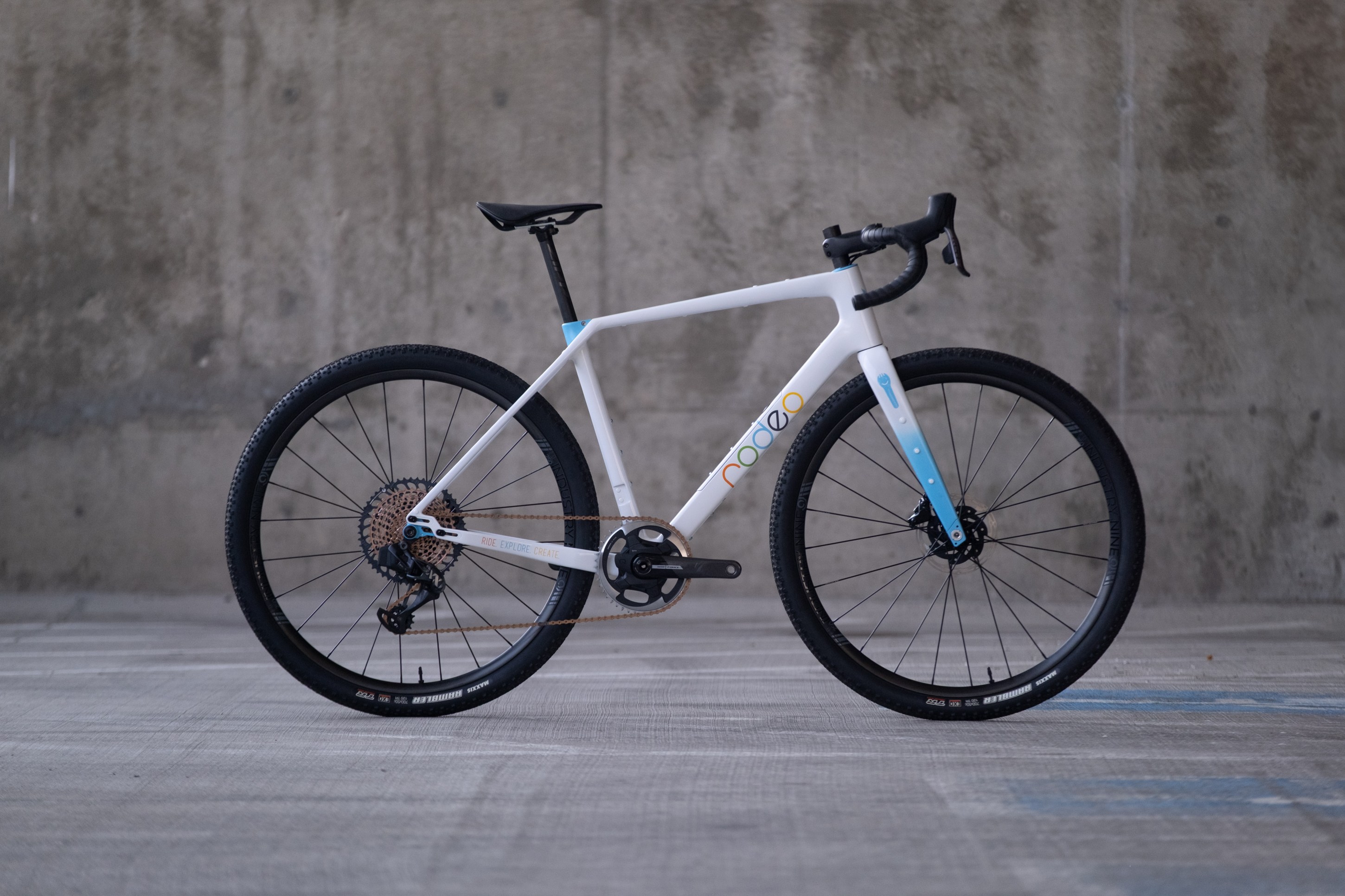 A modern white bicycle with blue accents, featuring a sleek frame, disc brakes, and knobby tires, positioned against a concrete wall.