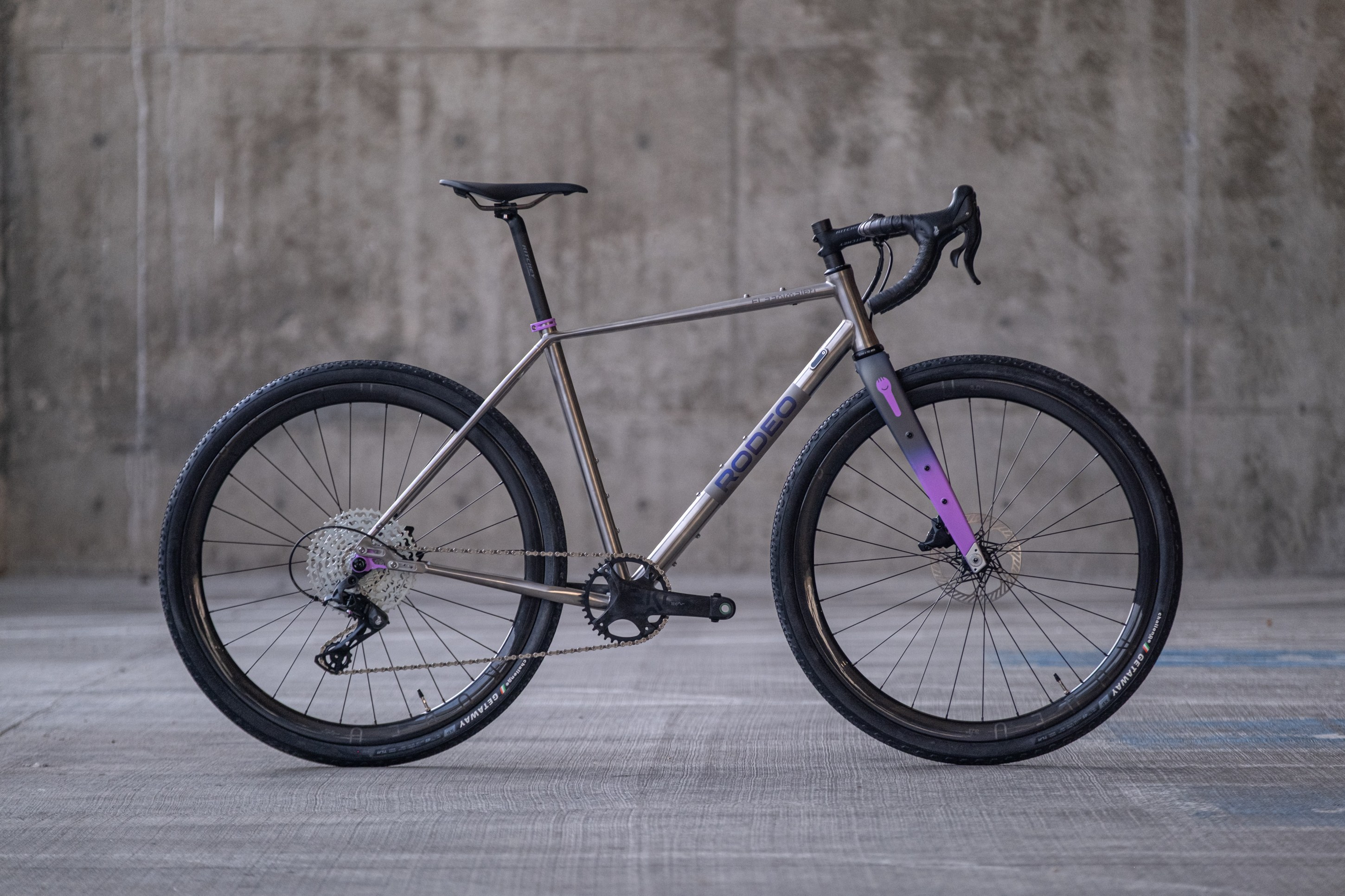 A modern gravel bike with a titanium frame and purple accents, posed against a concrete background.