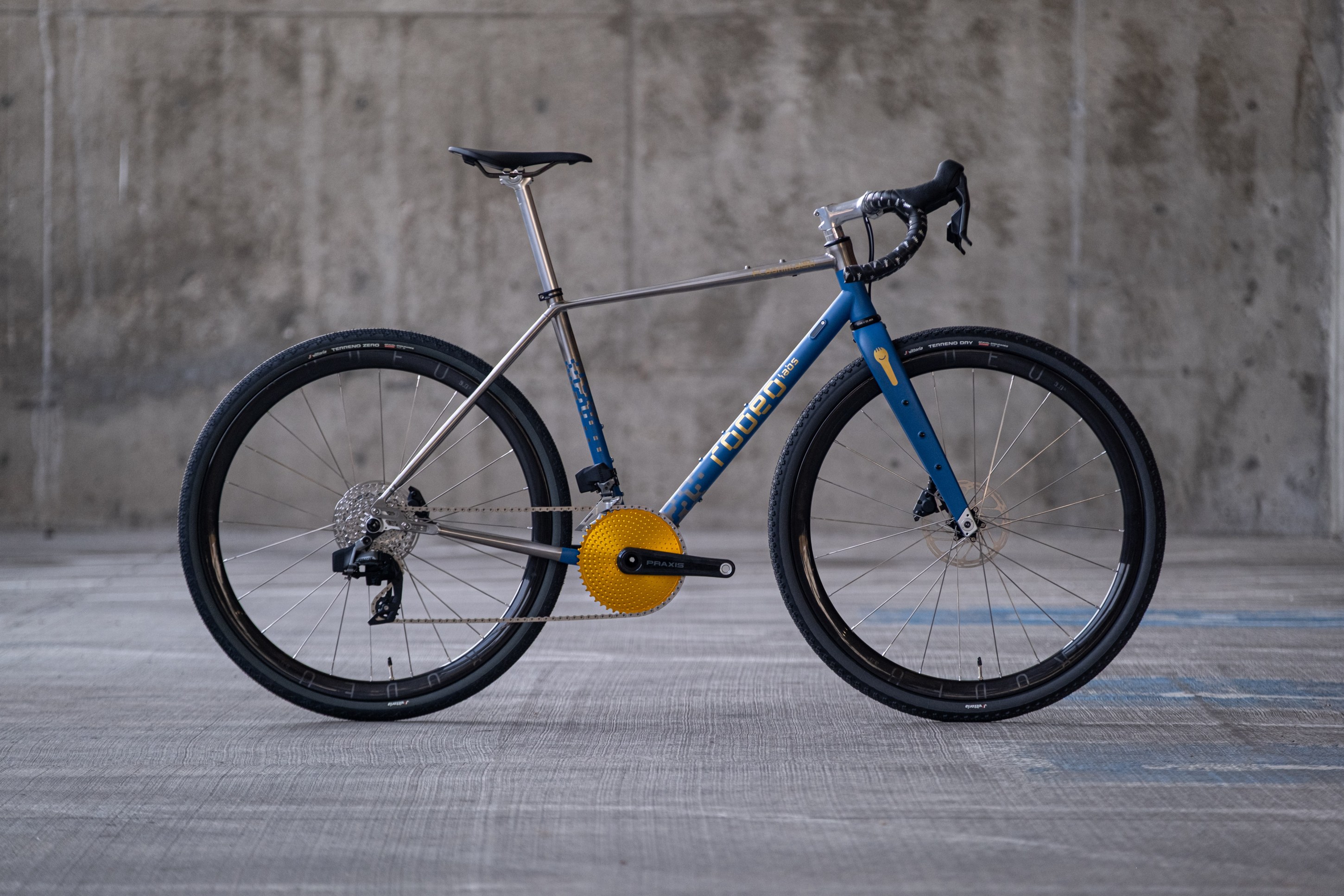A profile view of a modern bicycle with a blue and silver frame, black tires, and a distinctive yellow gear system, set against a concrete background.