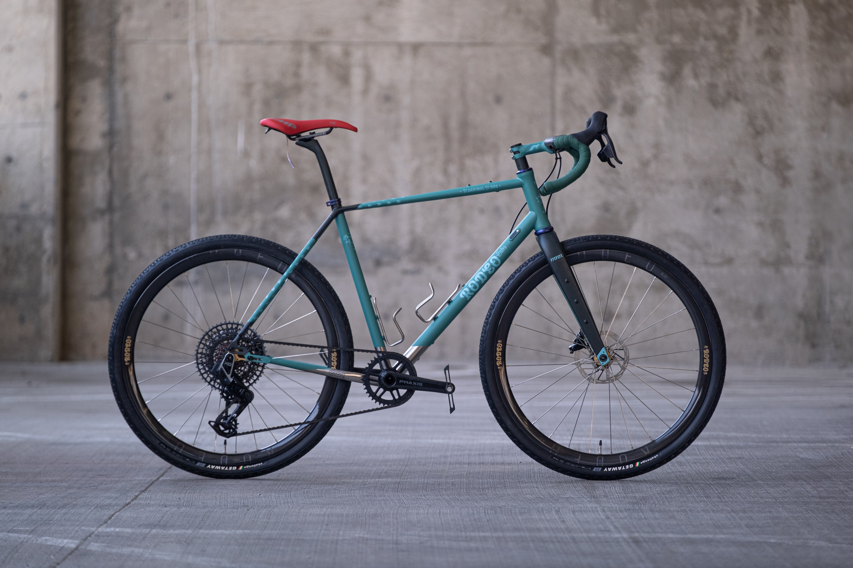 A turquoise gravel bike with a red saddle, disc brakes, and wide tires, positioned against a textured concrete wall.