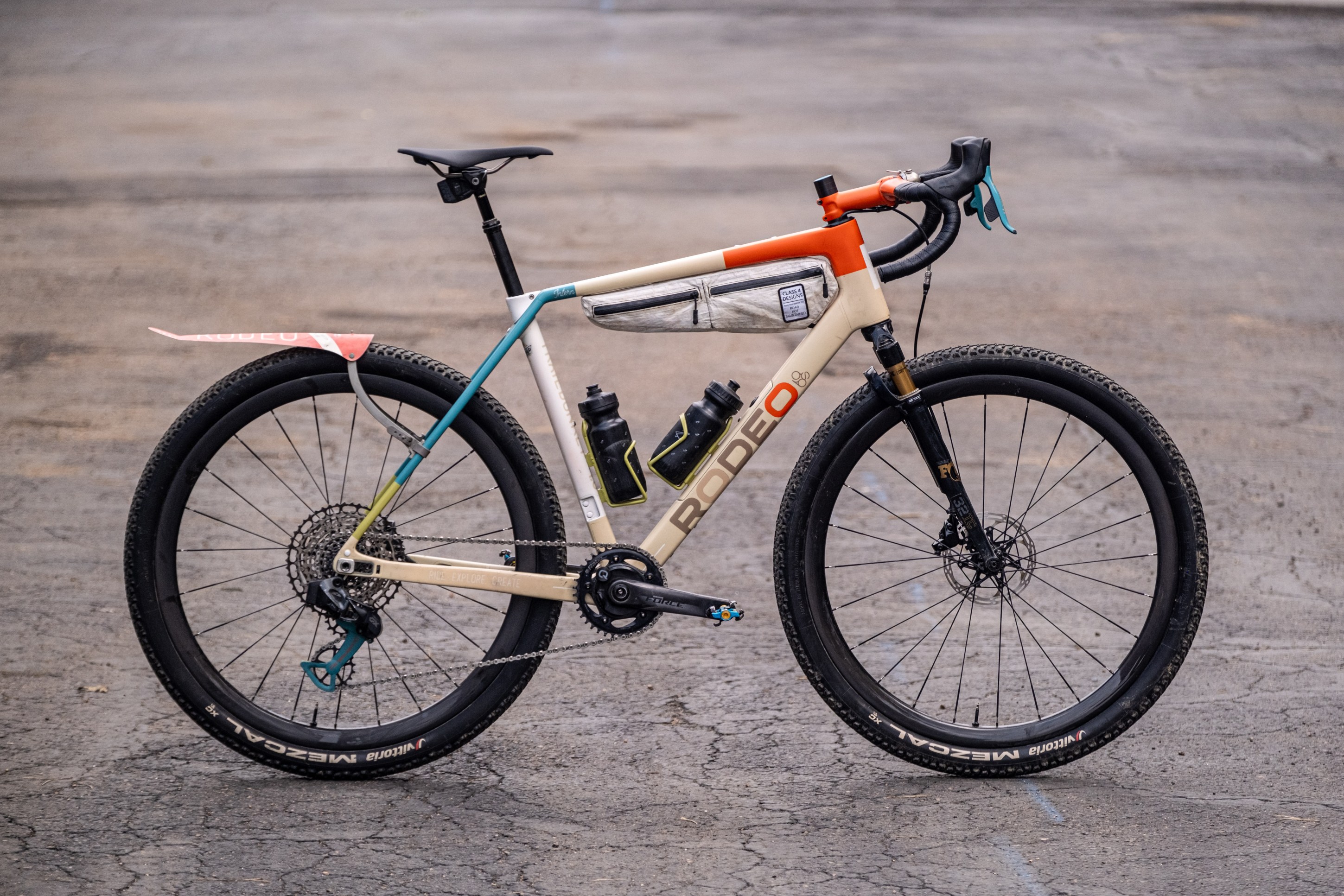 A stylish gravel bike with a cream and orange frame, featuring two water bottles, a front suspension, and wide tires, parked on an asphalt surface.