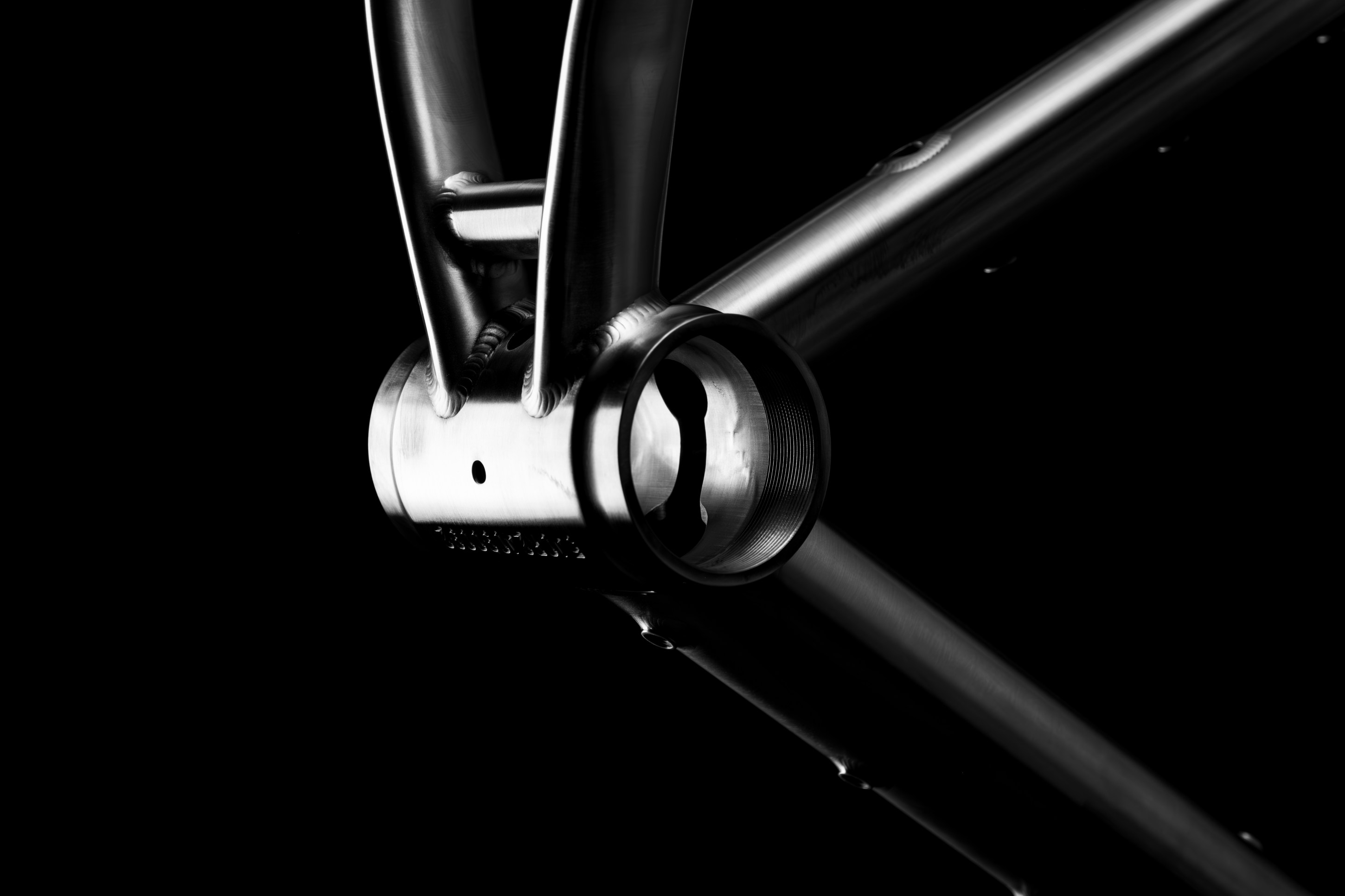 Close-up of a bicycle frame's bottom bracket area, showcasing polished metal and intricate welds against a black background.