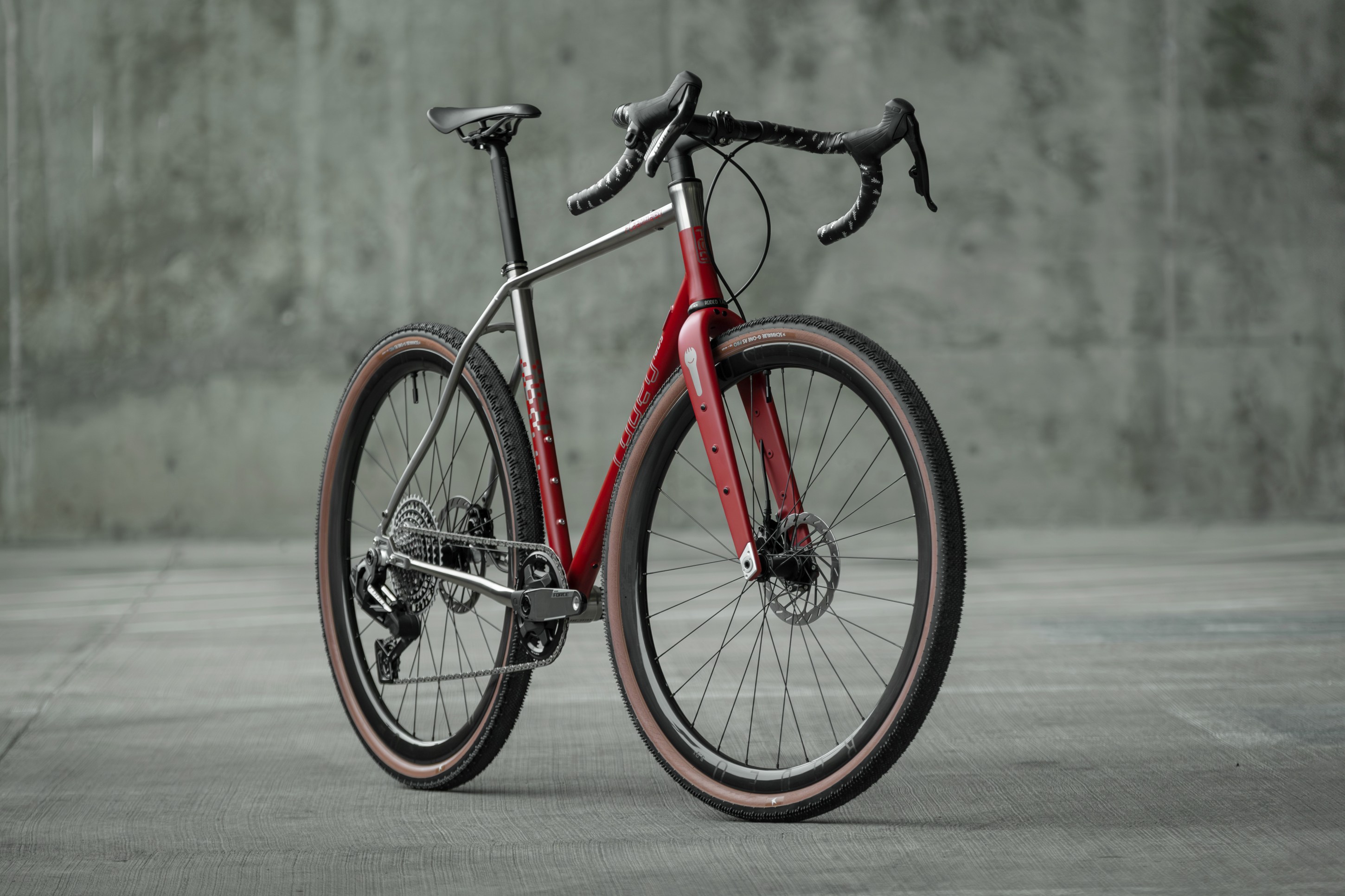 A modern bicycle with a sleek design, featuring a red and silver frame, drop handlebars, and wide tires, set against a textured gray background.