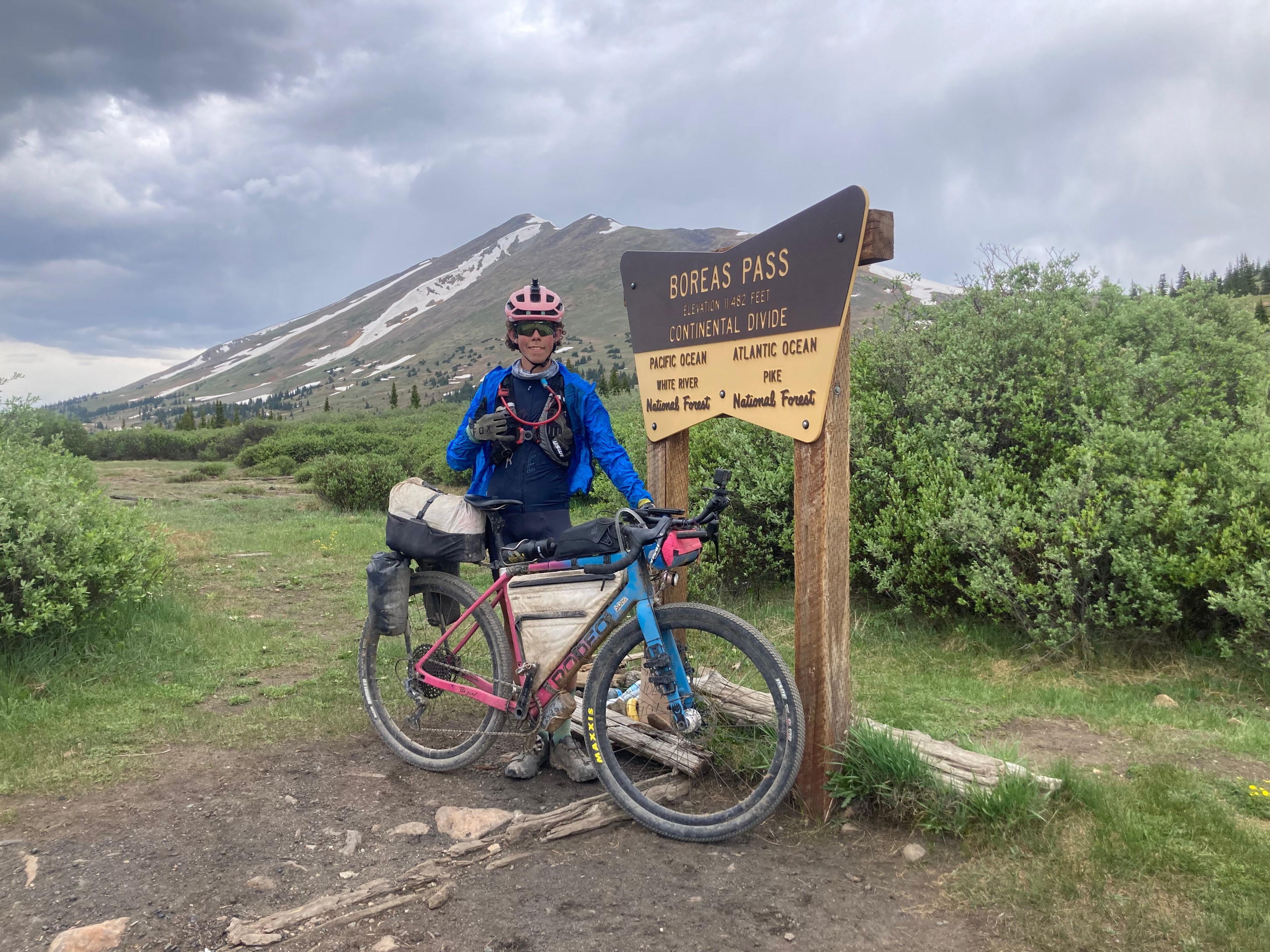 A cyclist stands beside a sign at Boreas Pass, marking the Continental Divide. The cyclist is wearing a helmet and blue jacket, with a fully loaded bike in the foreground. Snow-capped mountains are visible in the background under a cloudy sky.