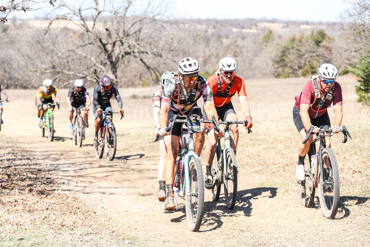 A group of six cyclists riding on a dirt path through an open landscape, with trees in the background. The riders are wearing colorful jerseys and helmets, engaged in a cycling event on a sunny day.