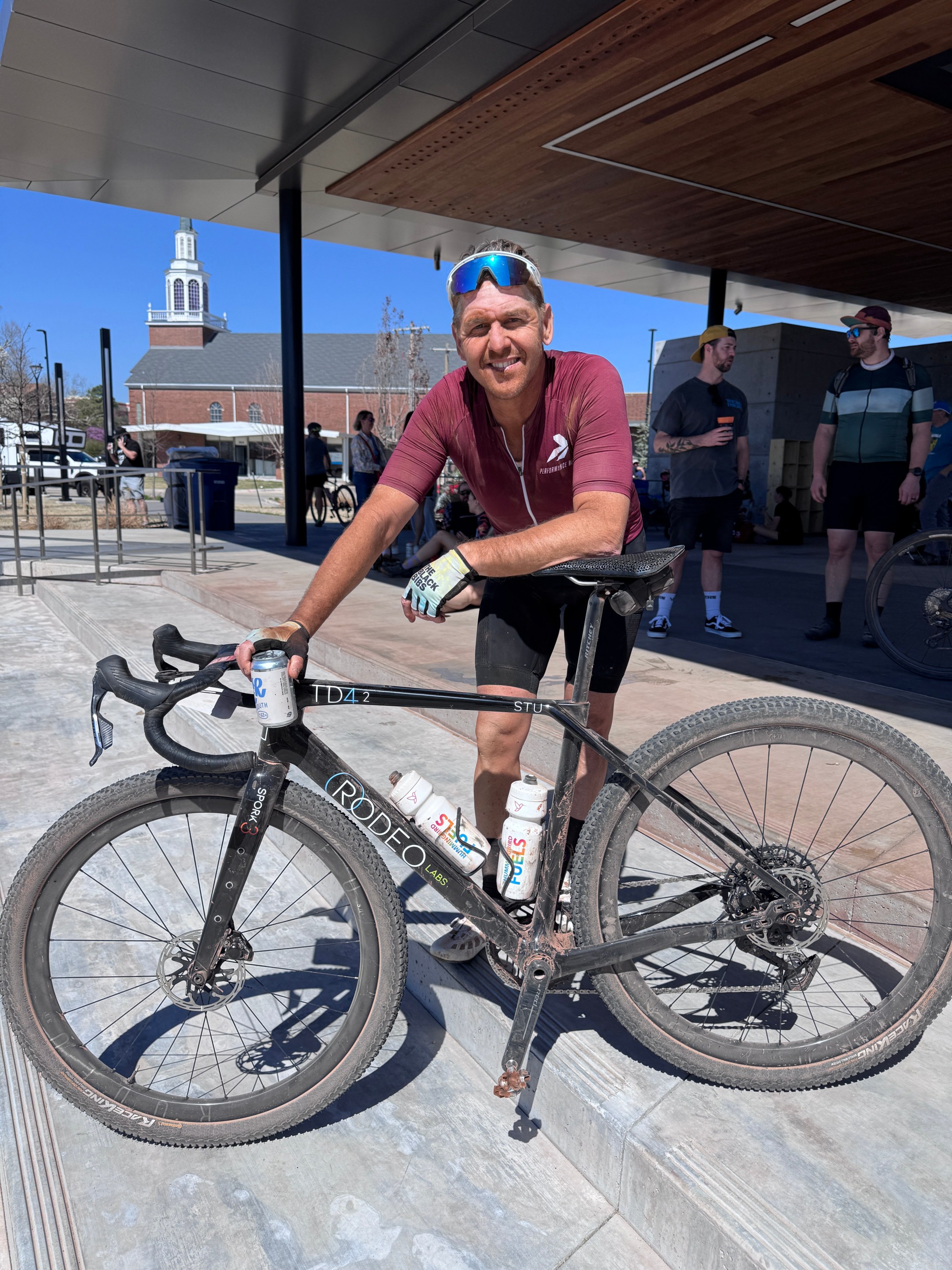 A cyclist in a maroon shirt smiles while holding a black bicycle with muddy tires in an outdoor setting, featuring a modern building in the background.