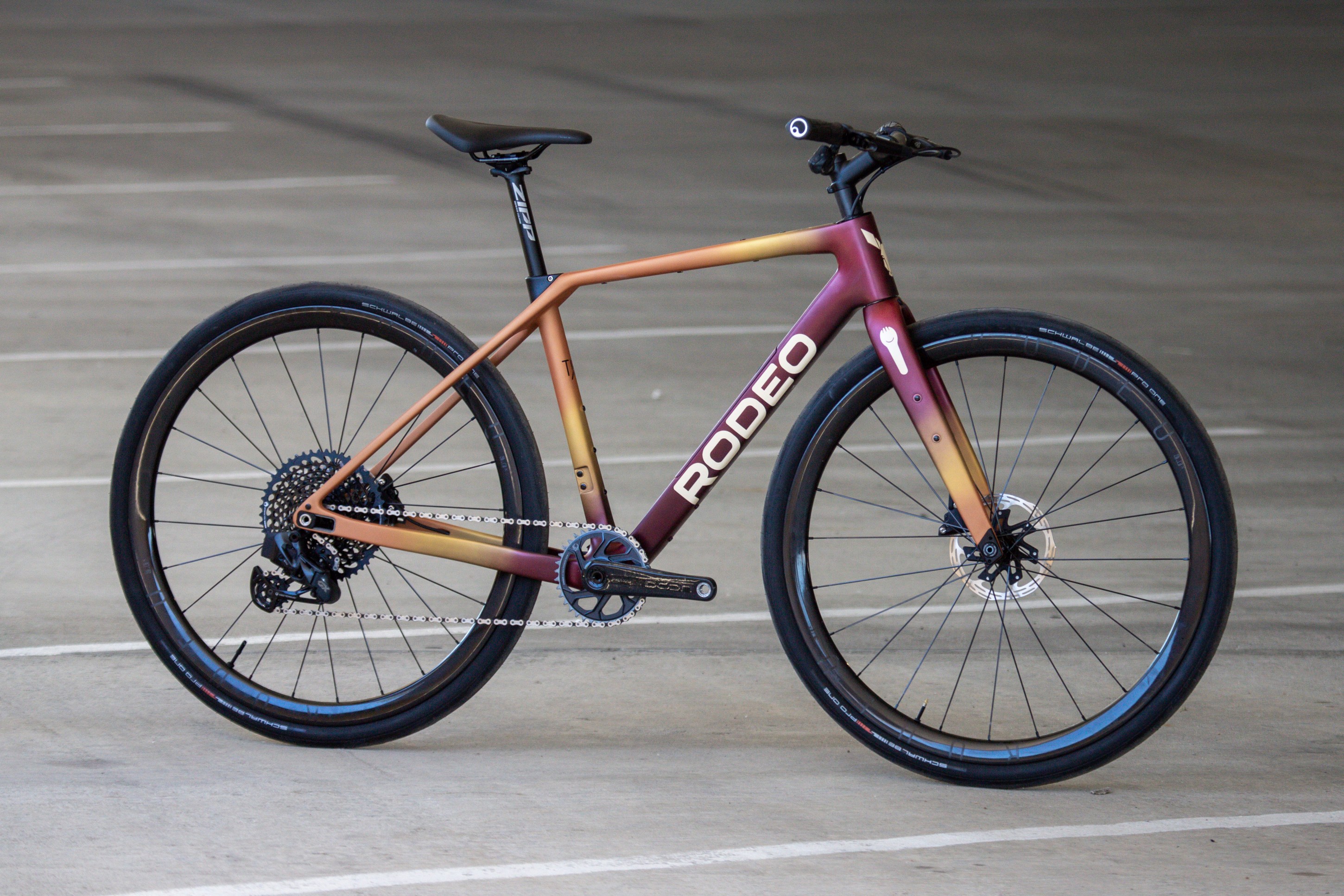 A modern bicycle with a gradient color design transitioning from orange to purple, featuring a sleek frame, disc brakes, and a single chainring setup, displayed on a concrete surface.