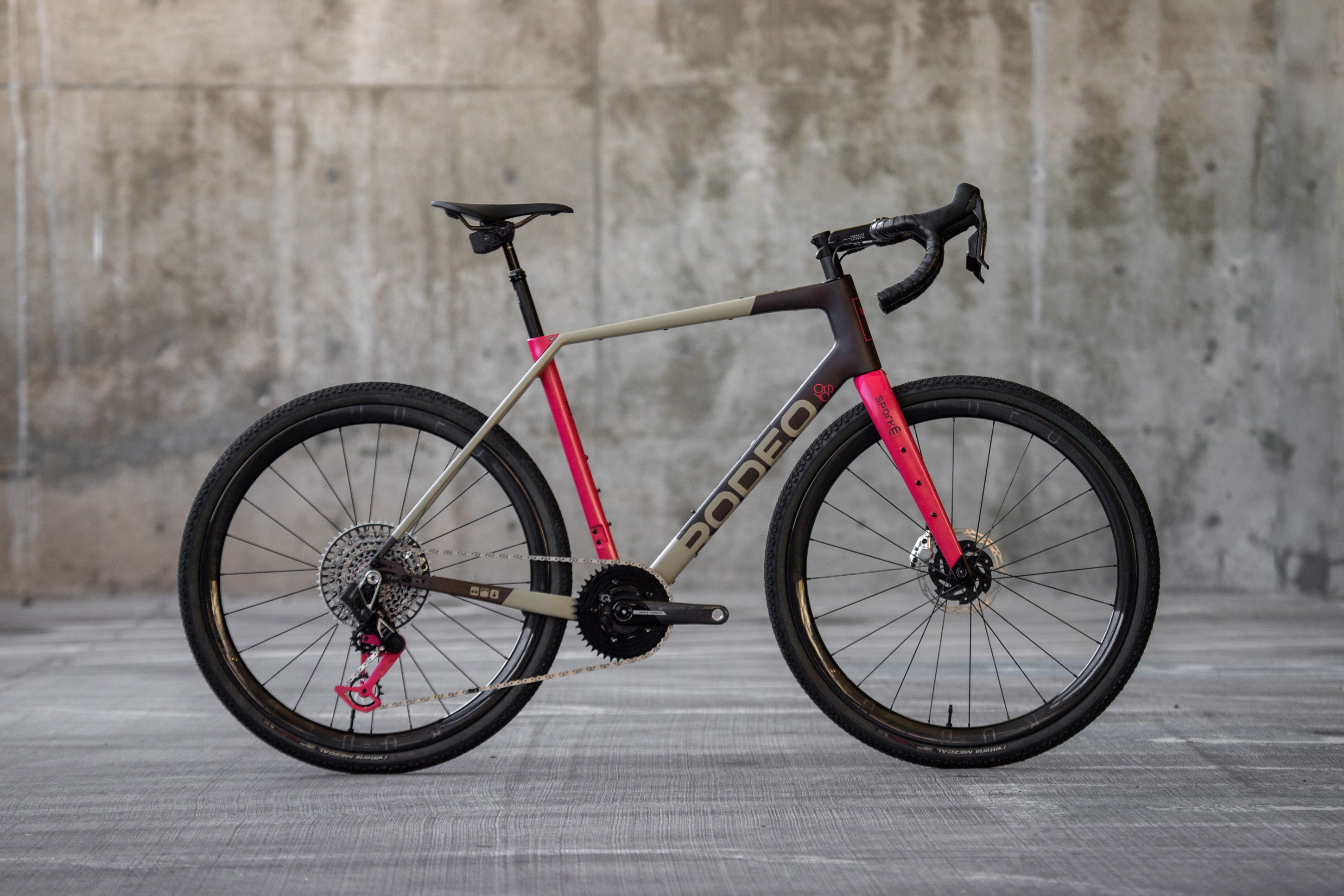 A modern gravel bike with a sleek design, featuring a gray and pink frame, disc brakes, and wide tires, set against a concrete background.