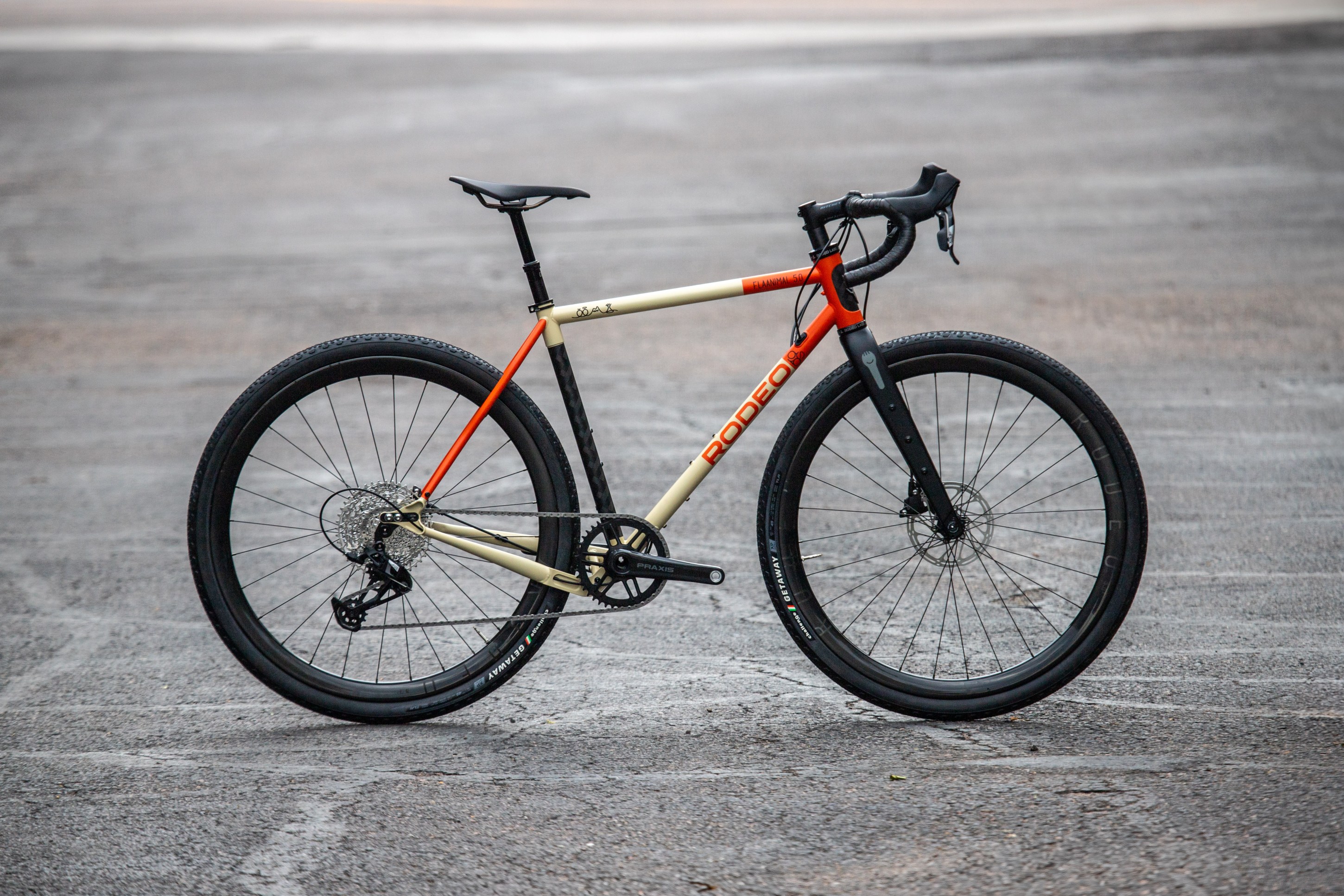 A modern gravel bike with a tan and orange frame, featuring disc brakes and wide tires, positioned on a gray asphalt surface.