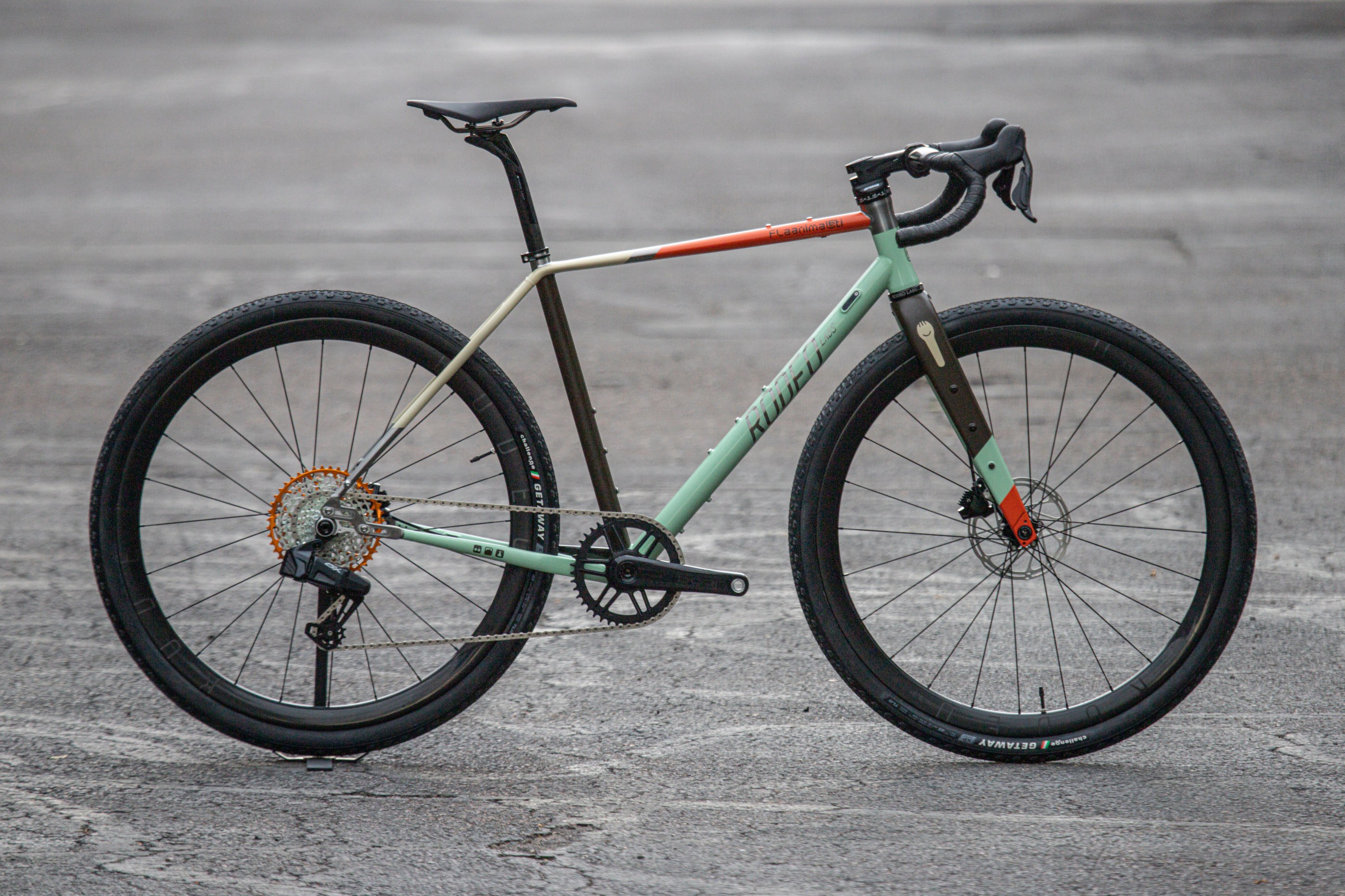 A modern gravel bike with a mint green and orange color scheme, featuring large black tires and a sleek design.