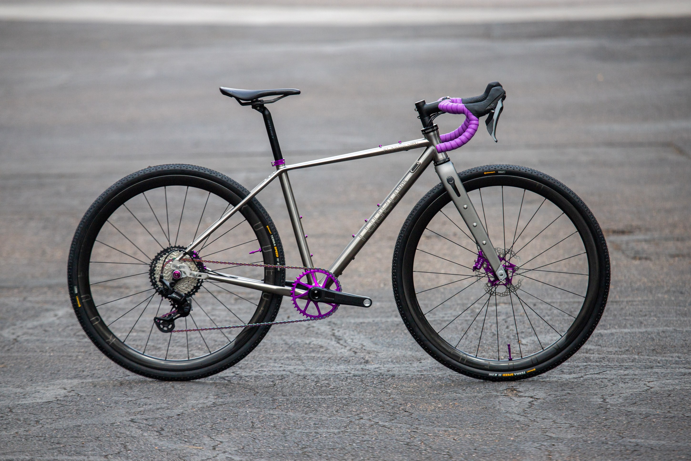 A modern bicycle with a silver frame and vibrant purple accents, showcasing a single front chainring and wide tires, set against a textured surface.