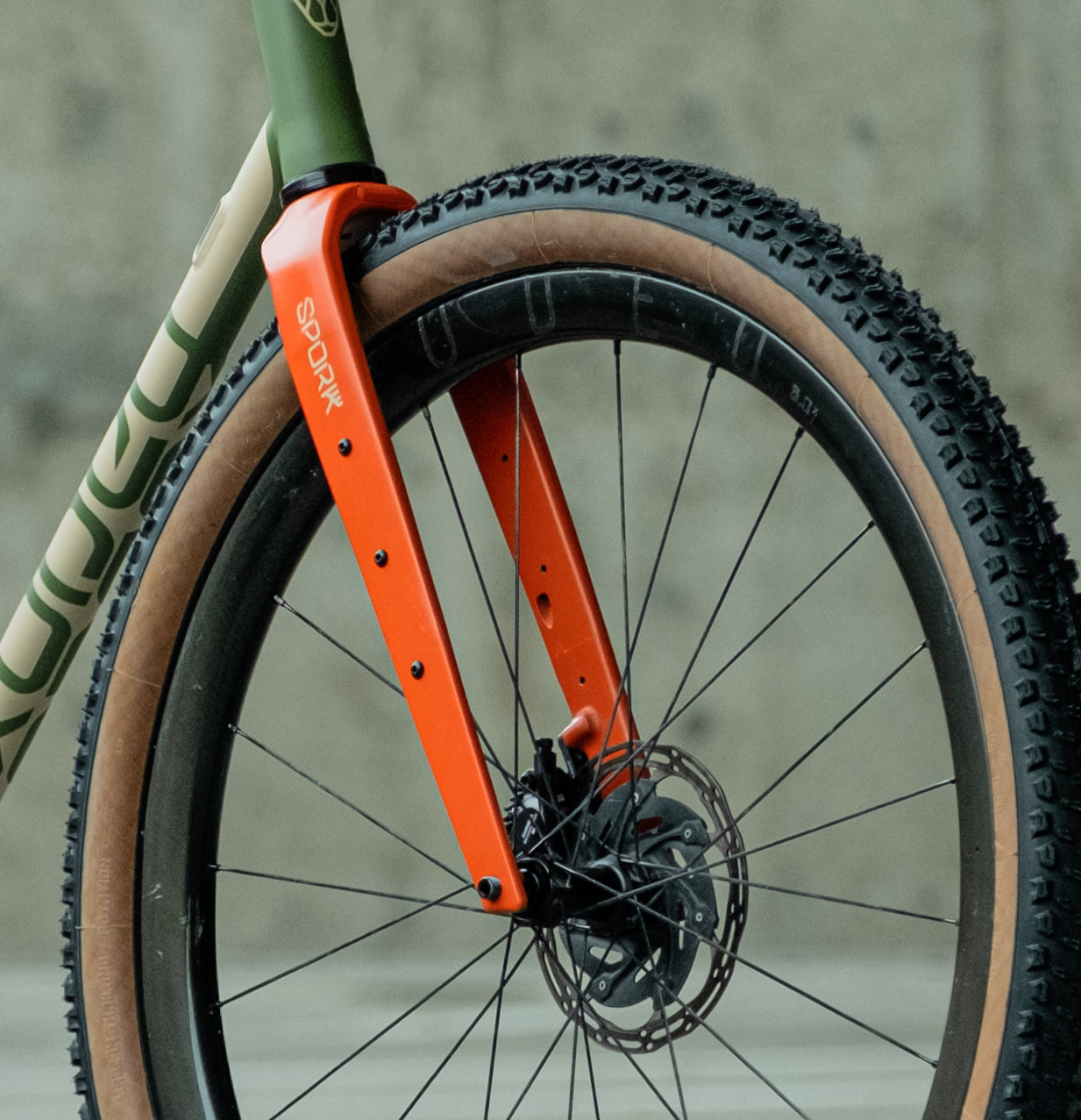 Close-up of a bicycle front fork featuring an orange color and disc brake, paired with a knobby tire and a partially visible green bike frame.
