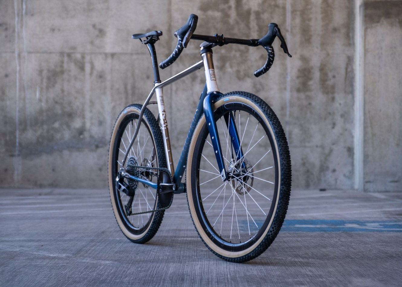 A modern gravel bike with a silver and blue frame, featuring drop handlebars and wide tires, positioned in an industrial setting with concrete background.
