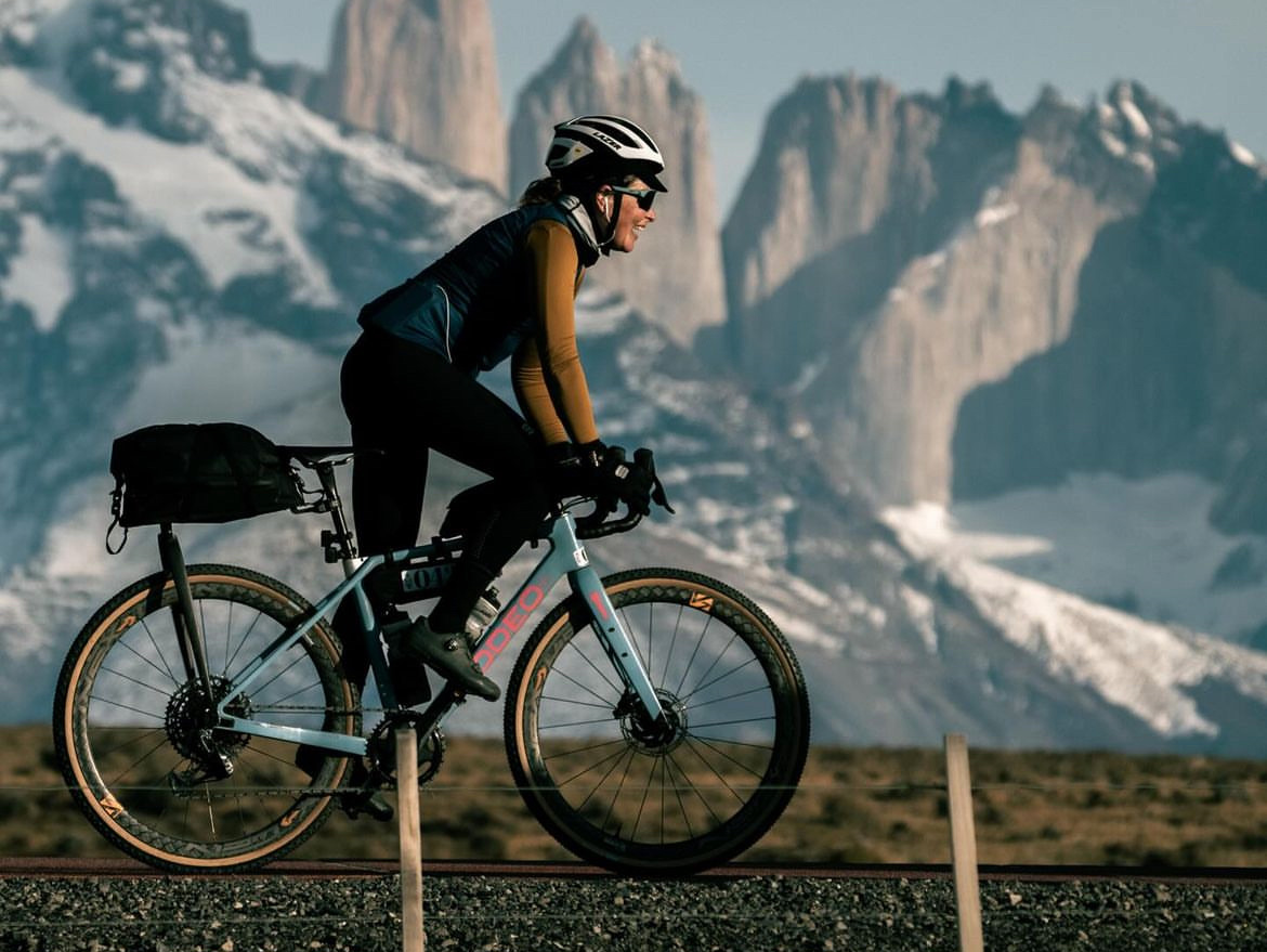 A cyclist riding a blue gravel bike along a road with a mountainous landscape in the background, featuring snow-capped peaks and a clear sky.