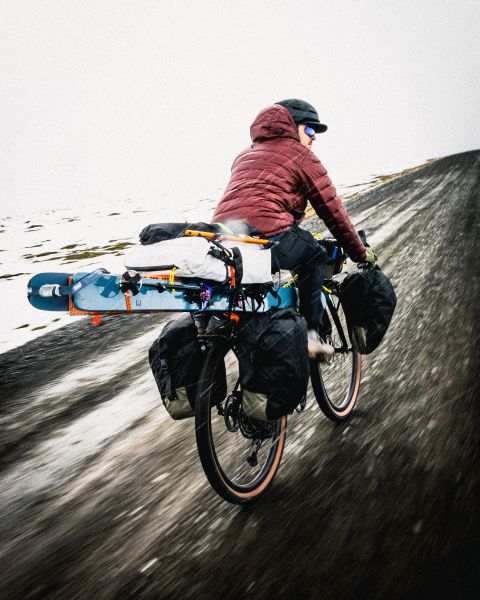 A cyclist riding on a rugged path, carrying a snowboard and equipped with various bags.