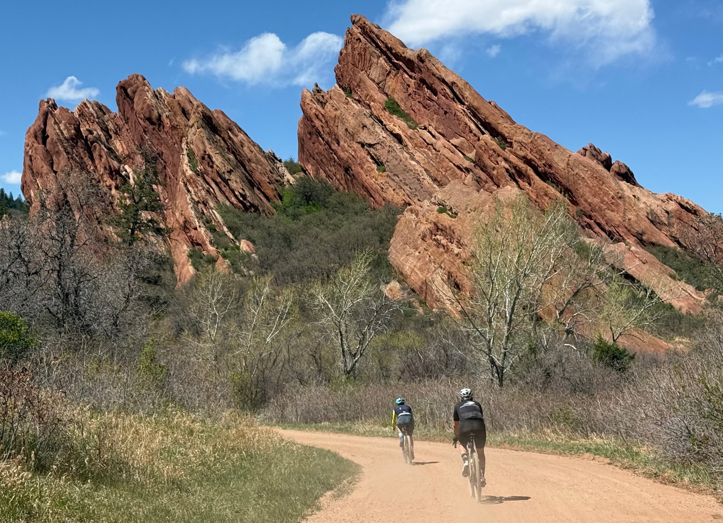 Two cyclists riding on a dirt path with striking red rock formations in the background under a blue sky.