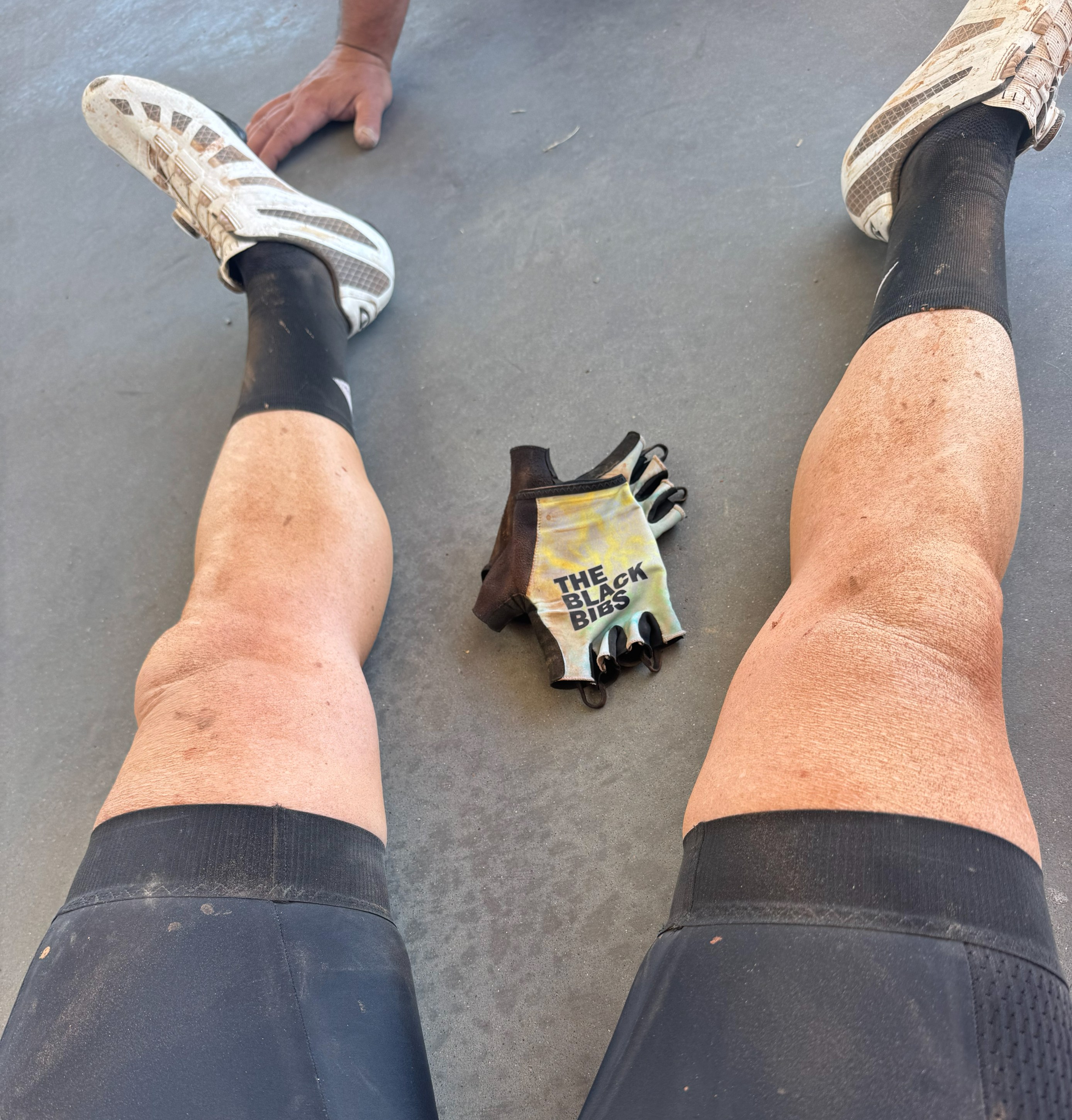 Two legs resting on a gray surface, wearing black cycling shorts and white sports shoes, with a pair of dirt-smudged gloves featuring the text 'THE BLACK BIBS' lying beside them.