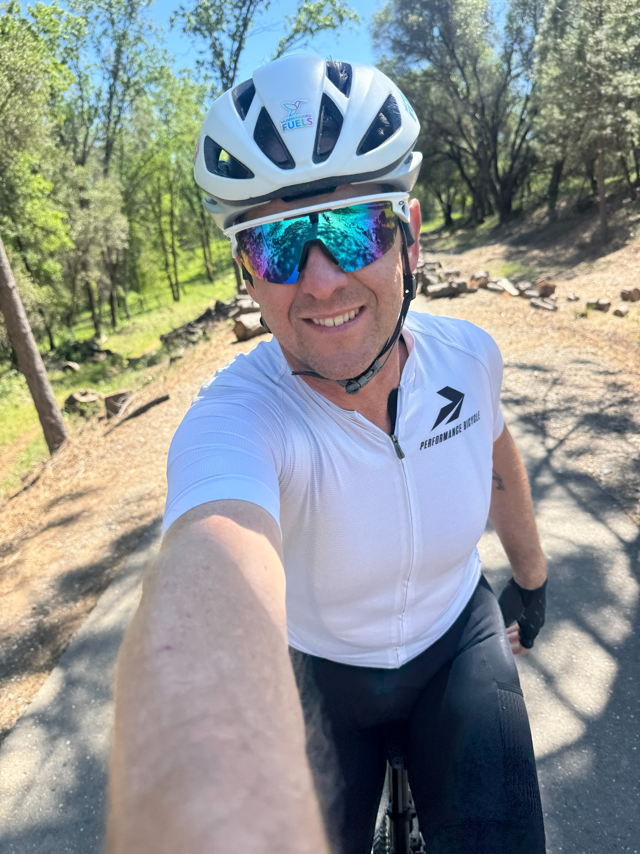 A cyclist wearing a white helmet and sunglasses, smiling while taking a selfie on a bike path surrounded by greenery.