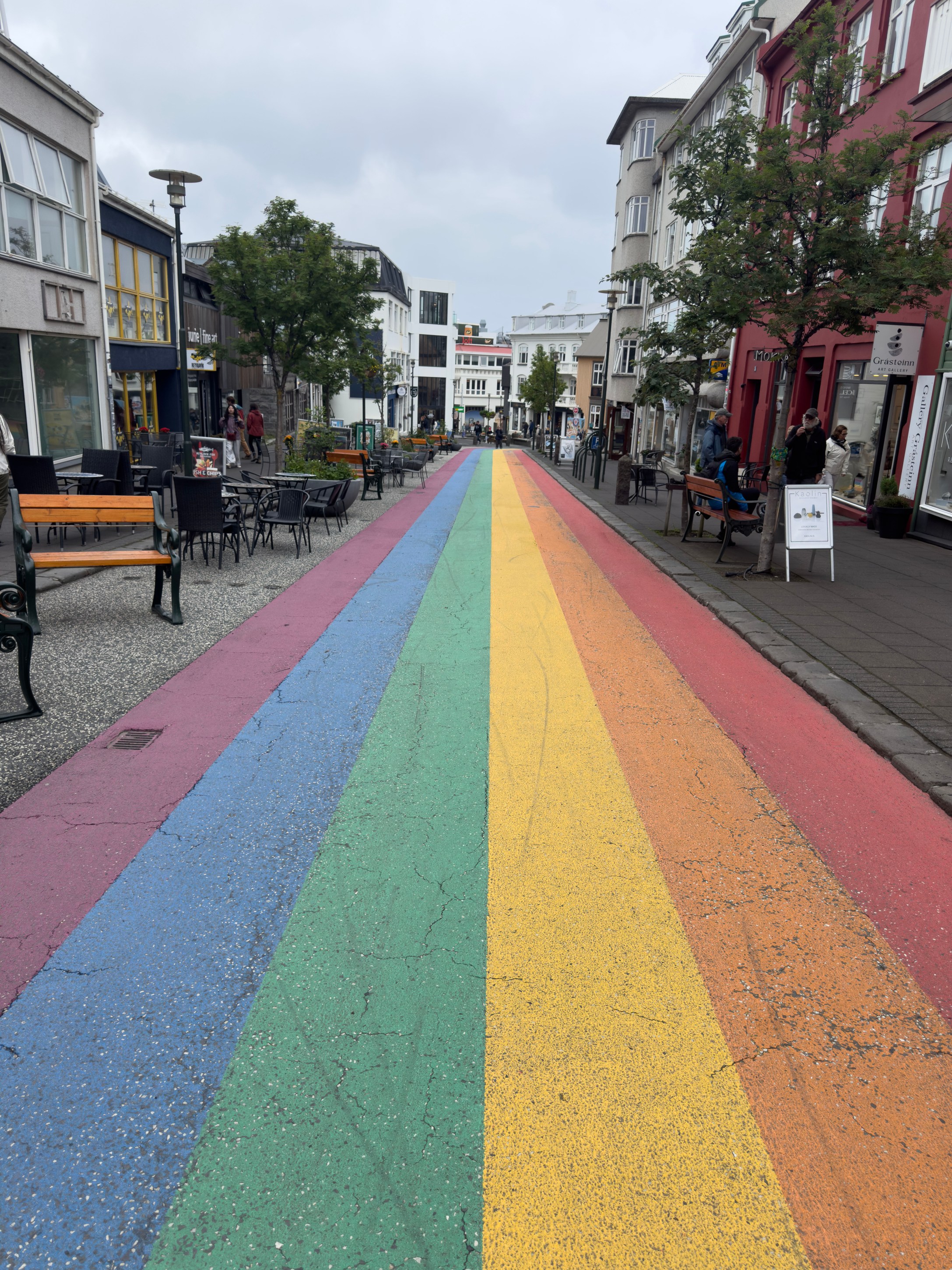 A colorful rainbow-painted street lined with outdoor seating and shops, under a cloudy sky.