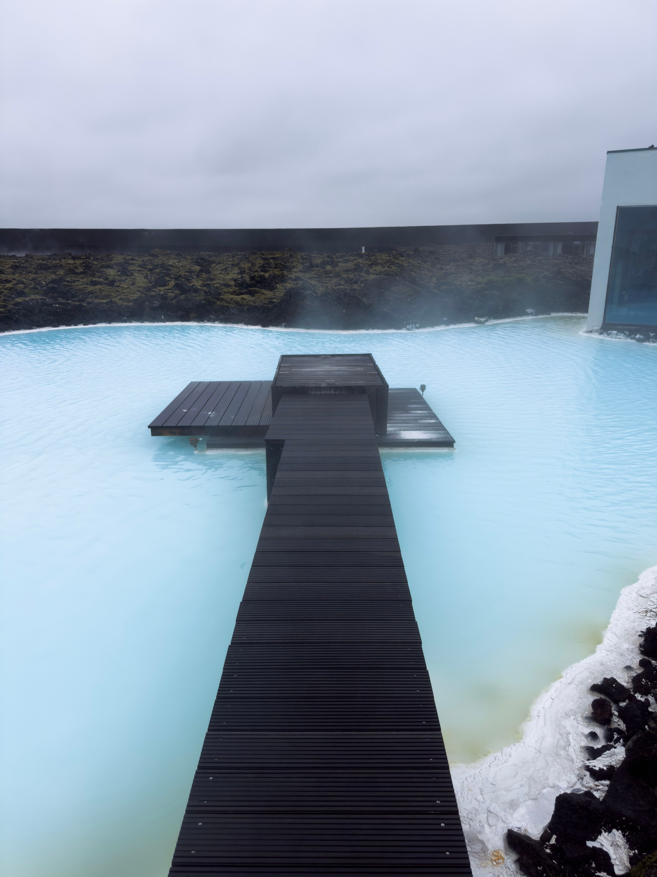 A wooden walkway extends over a tranquil, blue geothermal pool, surrounded by dark volcanic rock and white mineral deposits under a cloudy sky.
