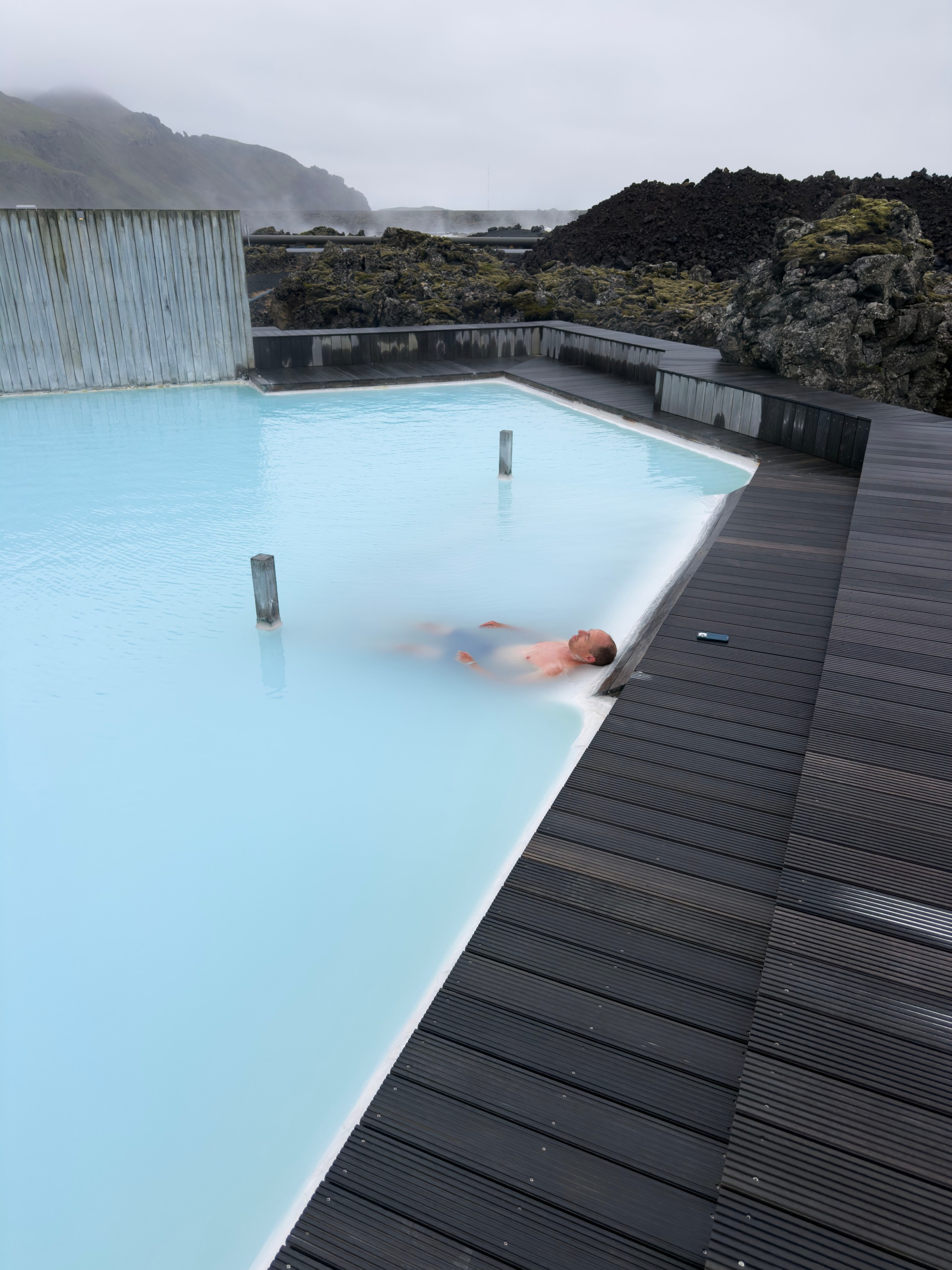 Person lounging in a blue geothermal pool with a rocky landscape and cloudy sky in the background.