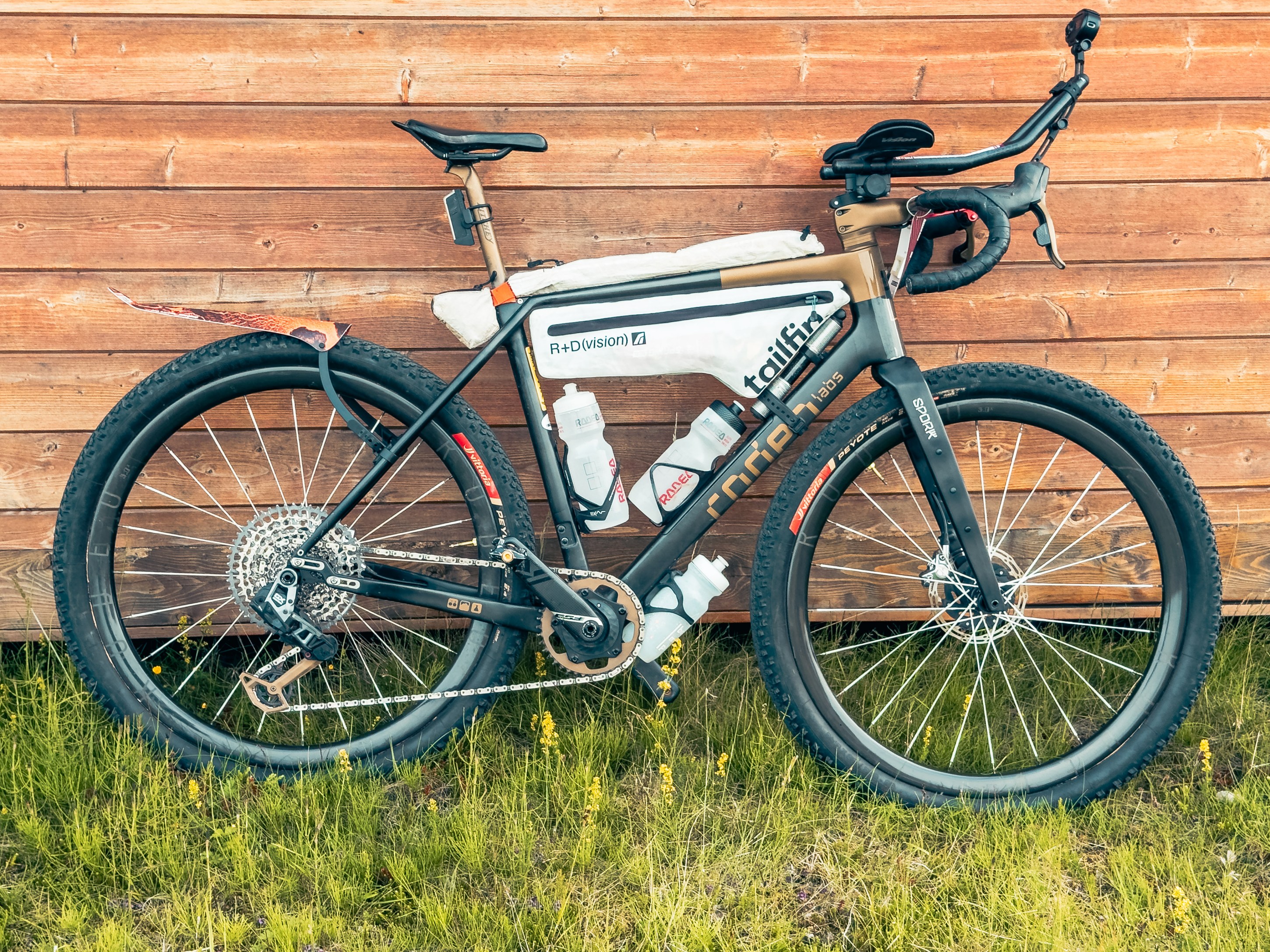 A gravel bike leaning against a wooden wall, featuring wide tires, a sleek frame, and equipped with water bottles and a saddlebag.