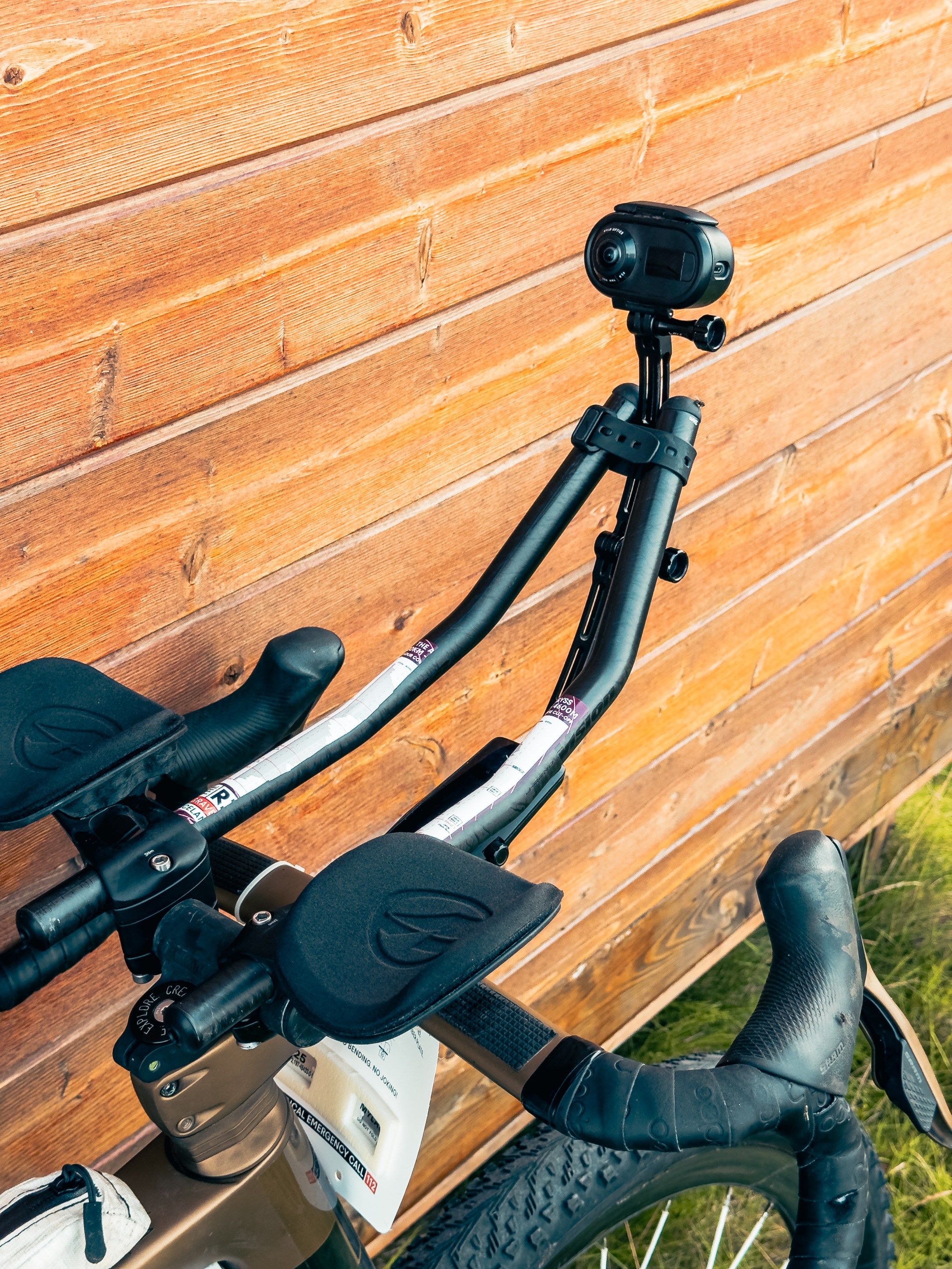 Close-up of bicycle handlebars featuring a camera mount, aerodynamic grips, and brake levers against a wooden background.