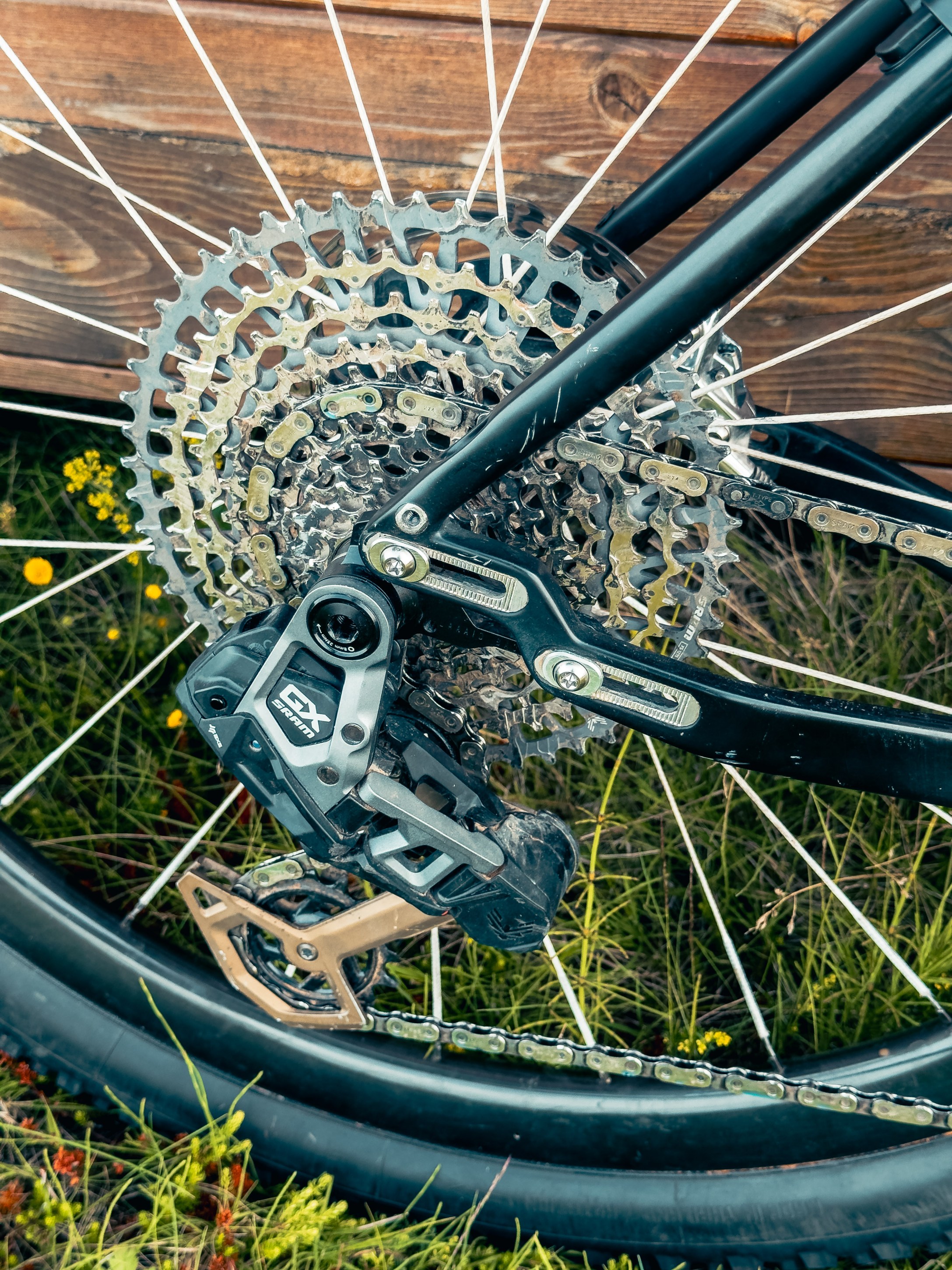 Close-up view of a bicycle's rear derailleur and cassette, showing the chain and gears against a background of grass and wooden surface.