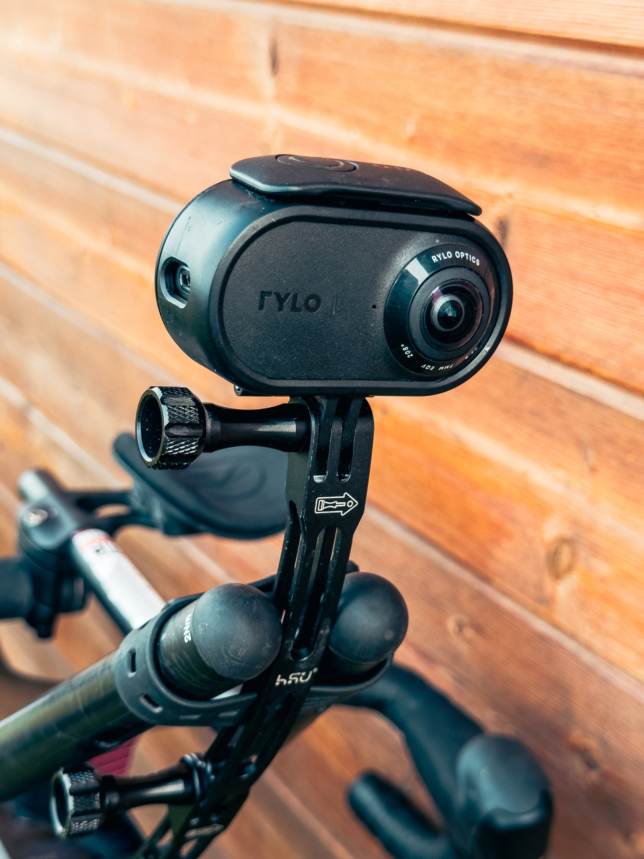 A close-up of a black Rylo camera mounted on a bicycle handlebar, set against a wooden background.
