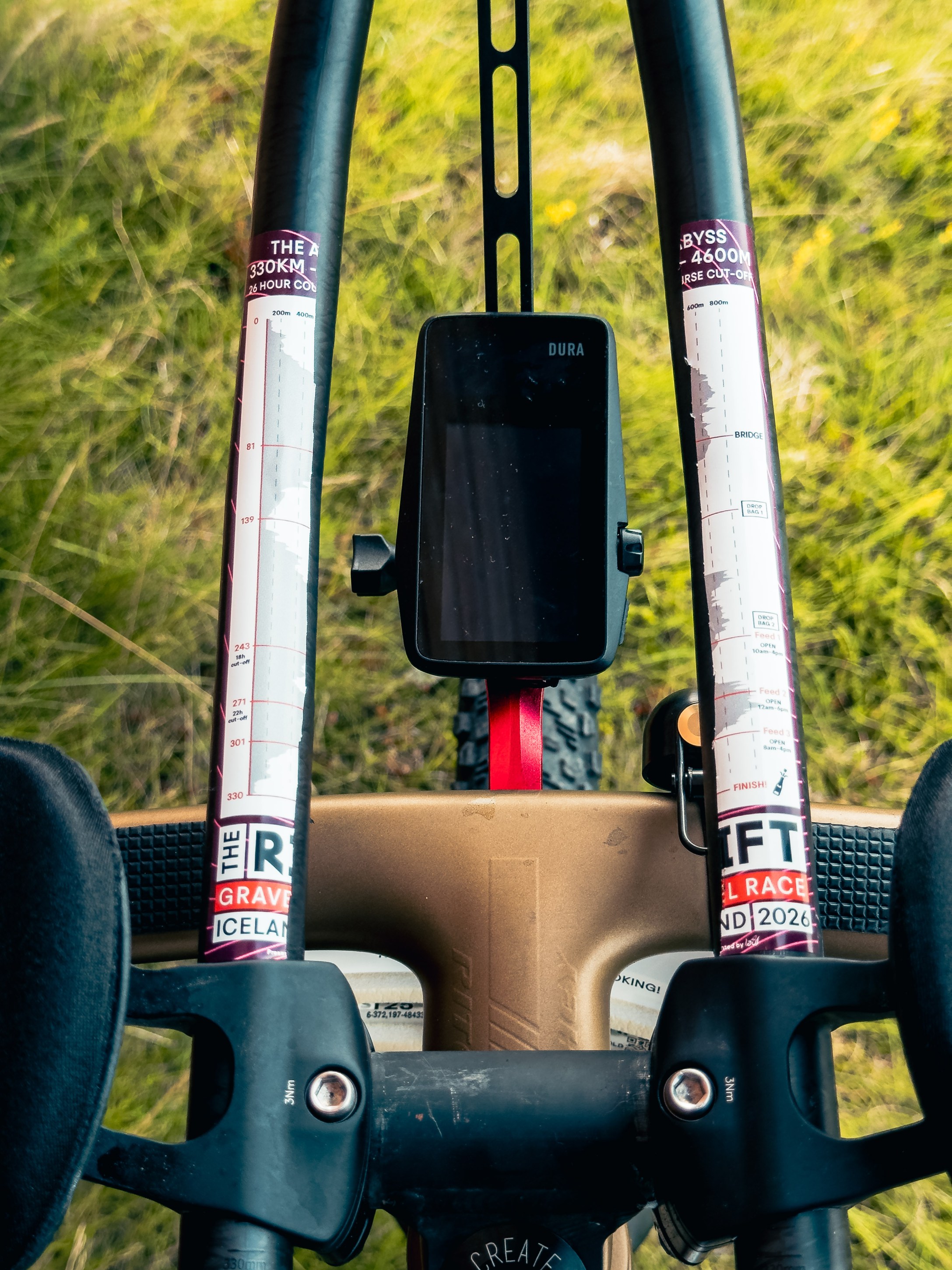 Close-up view of a gravel bike's handlebars featuring race distance markings and a GPS device displaying information against a blurred green background.
