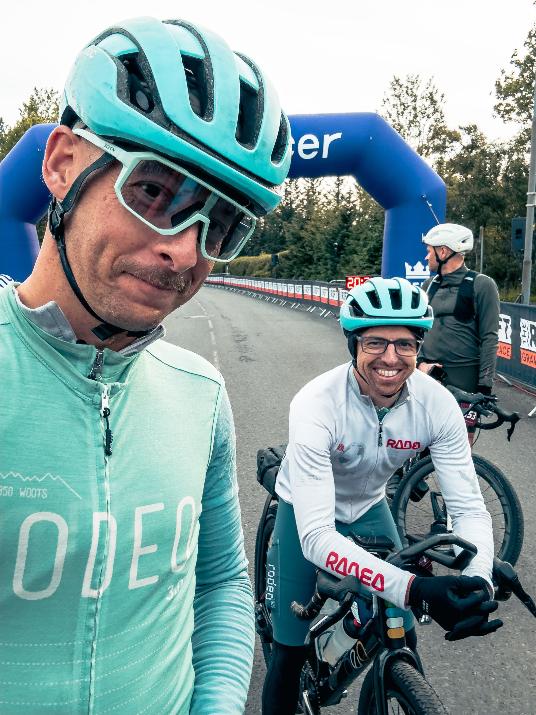 Two cyclists wearing helmets and cycling gear pose for a selfie at a cycling event, with an inflatable arch in the background.