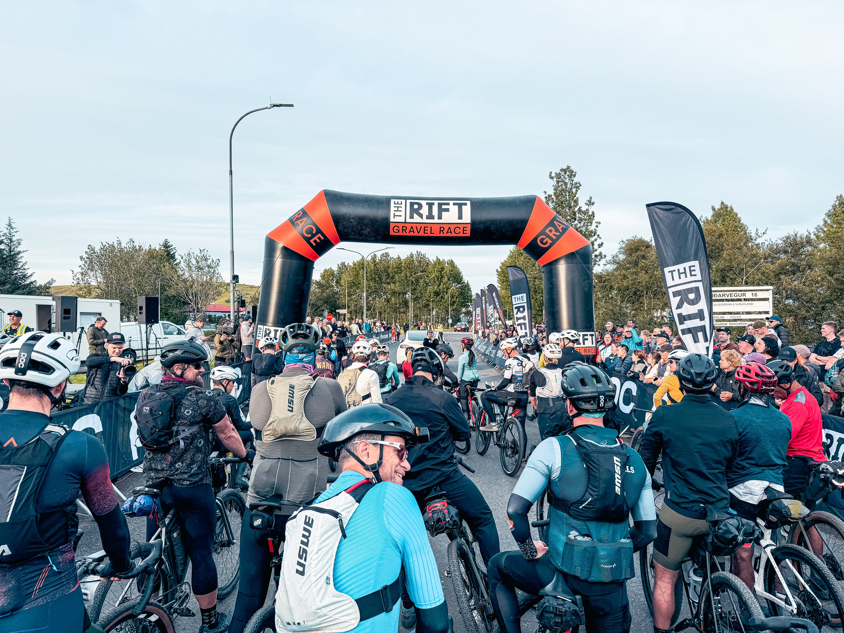 A crowd of cyclists in helmets and gear gathered near the starting line of the RIFT Gravel Race, with a large inflatable arch marking the event and spectators in the background.
