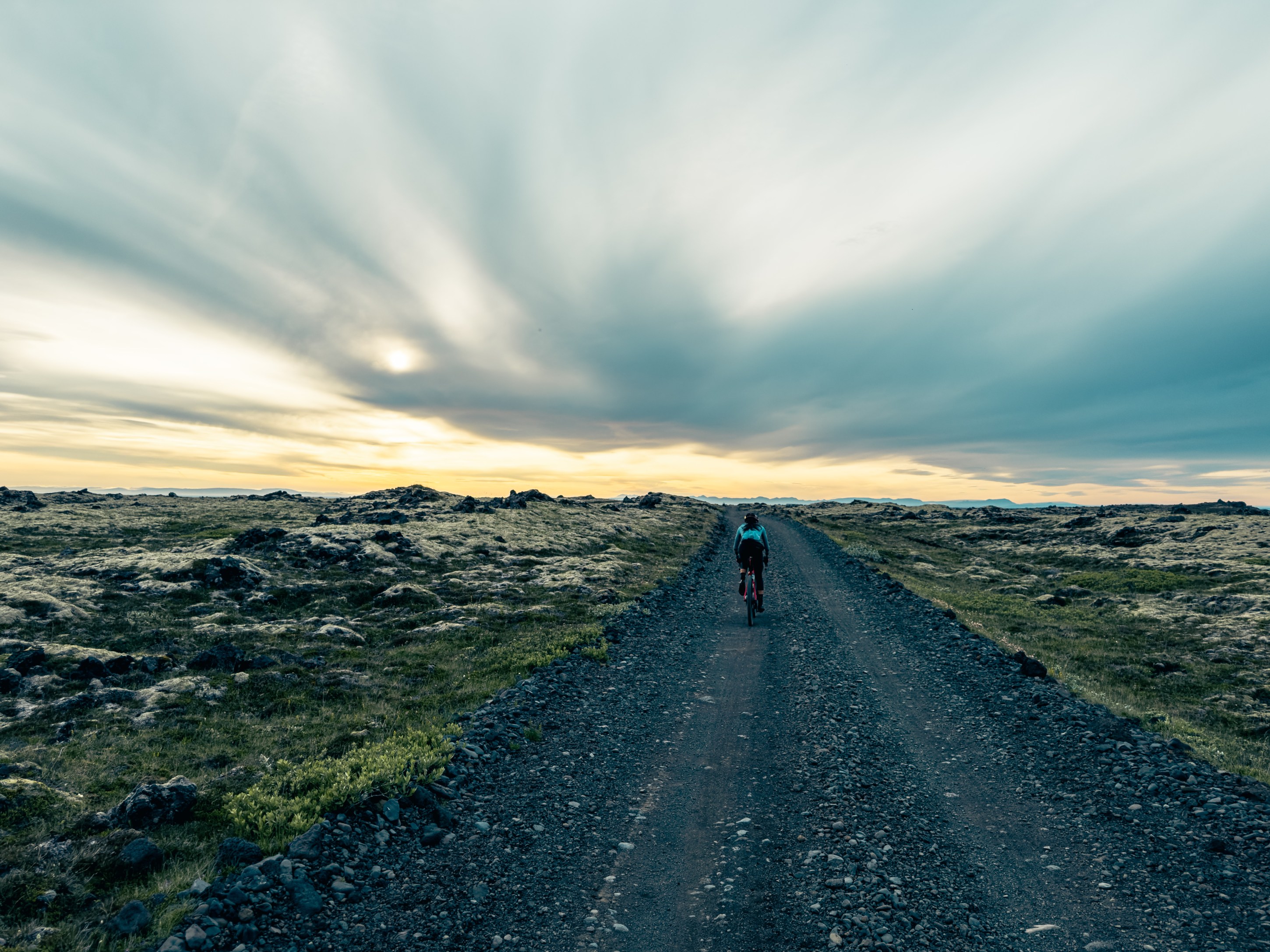 A cyclist riding along a rocky dirt road during sunset, with expansive views of grassy terrain and dramatic clouds in the sky.