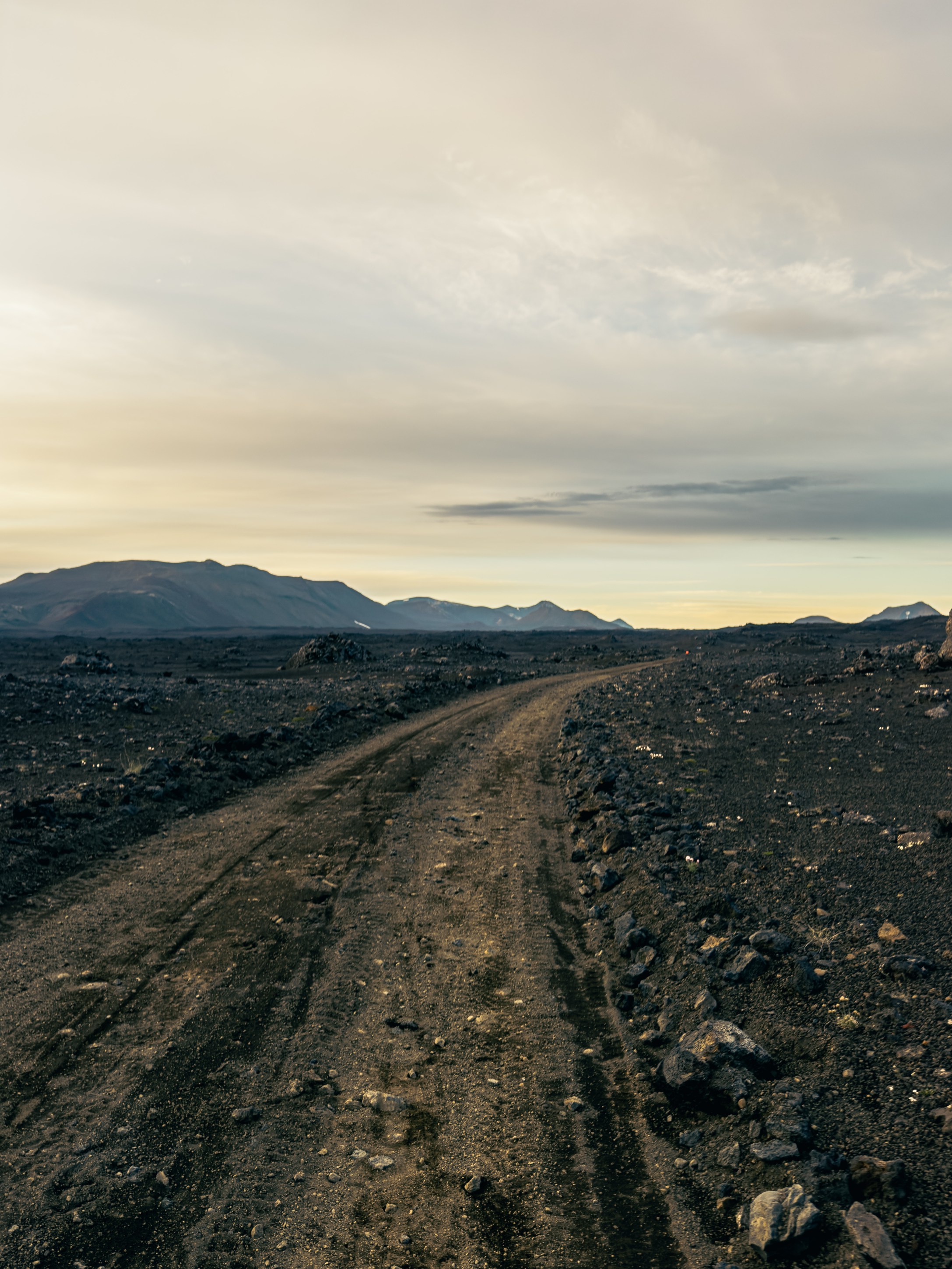 A winding dirt road cutting through a barren, rocky landscape with distant mountains and a cloudy sky.