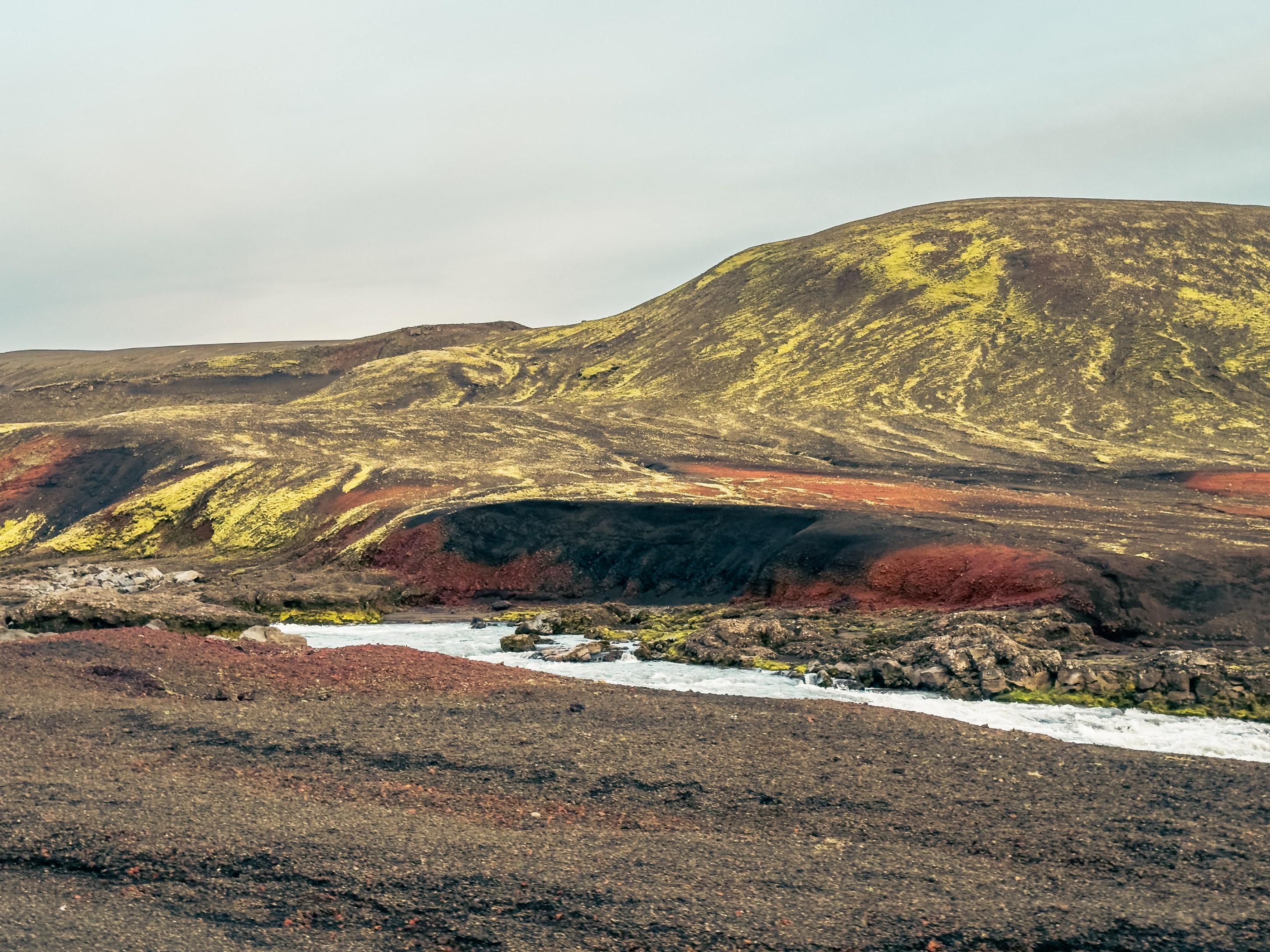 A landscape featuring volcanic hills with vibrant yellow, red, and black colored soil, alongside a flowing river in a rugged terrain.